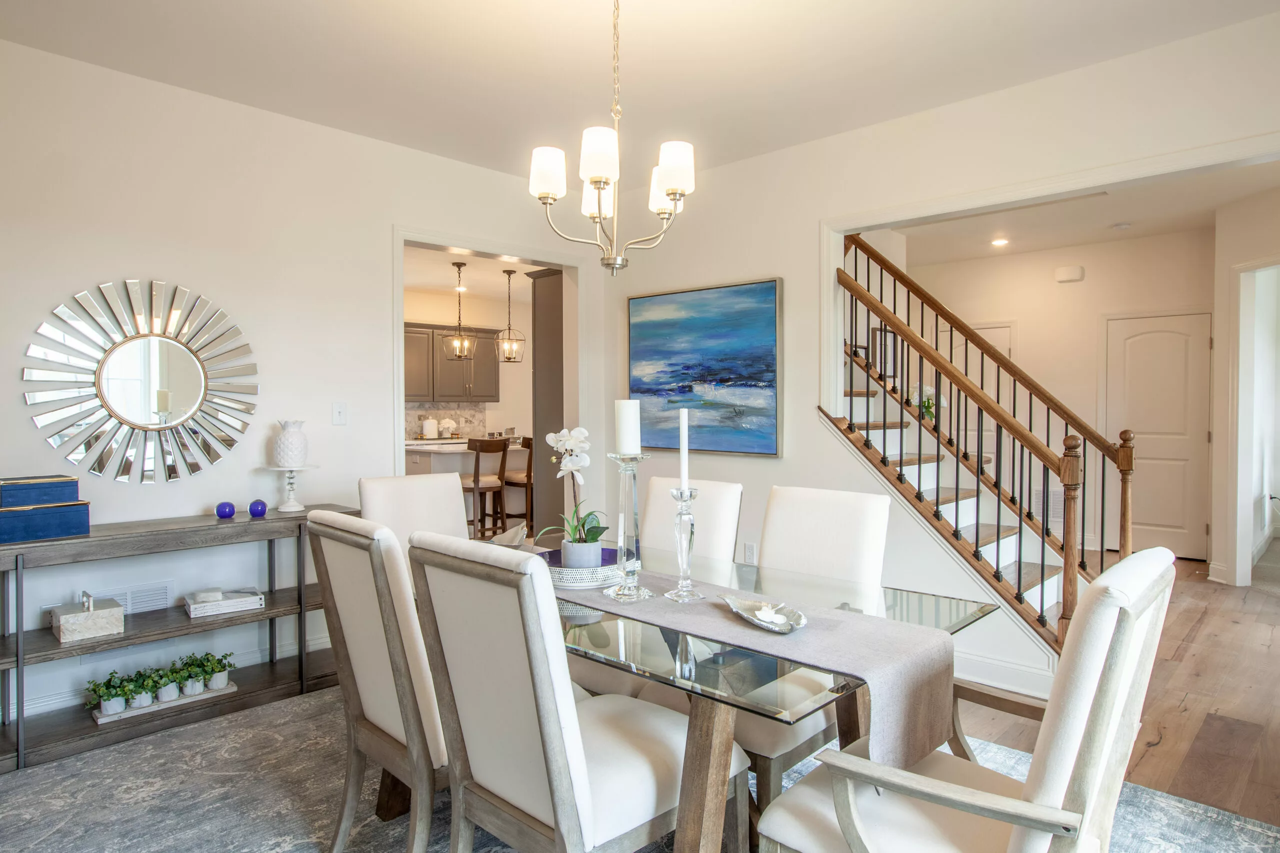 A modern dining room with a glass table set for six, cream upholstered chairs, a chandelier, wall art, and a decorative mirror. A staircase and a kitchen are visible in the background.