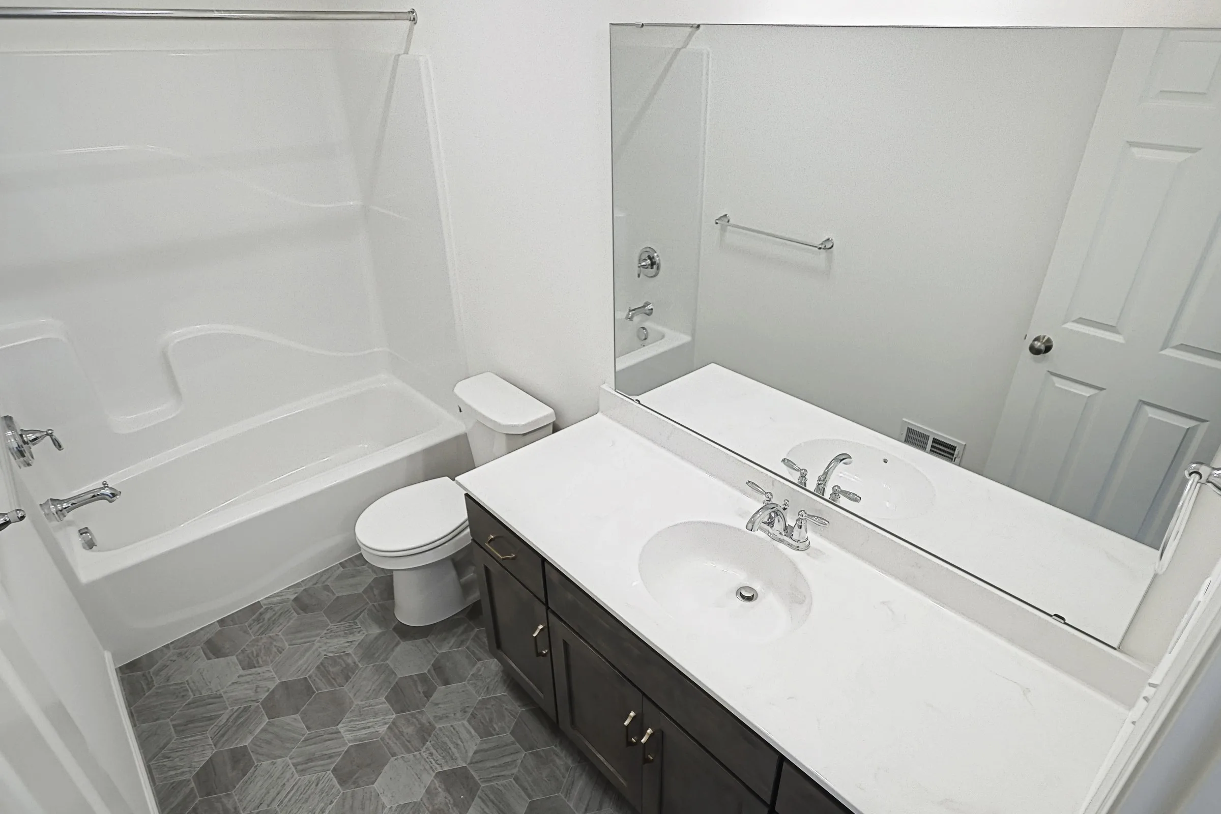 A modern bathroom with a white bathtub and shower, toilet, large mirror, and a dark vanity with a white countertop and sink. The floor has gray hexagonal tiles.