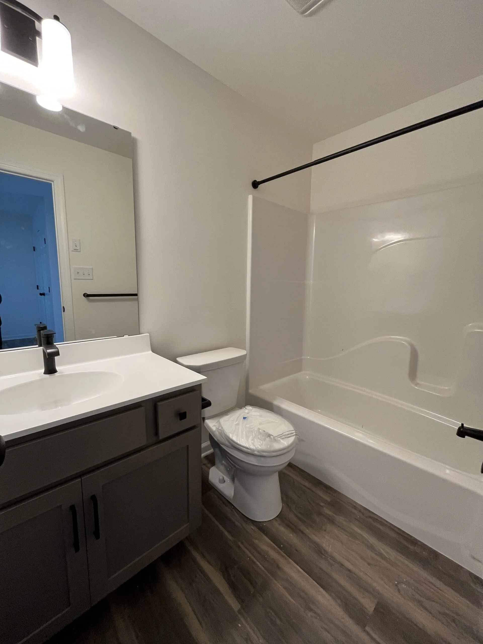 A small bathroom with dark wood flooring, a white bathtub-shower combo, a toilet with plastic wrap, a gray vanity with a white countertop, a large mirror, and a light fixture above the sink.