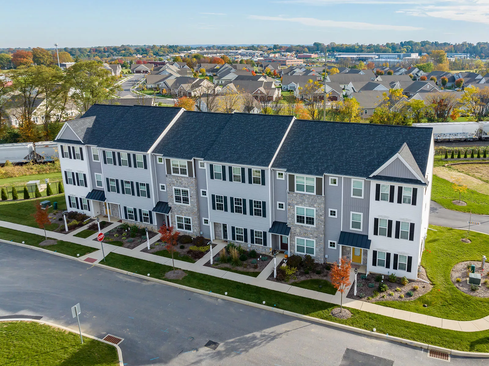 Aerial view of a row of modern three-story townhouses with black roofs, white and gray exteriors, and small landscaped front yards in a suburban neighborhood with trees and roads.