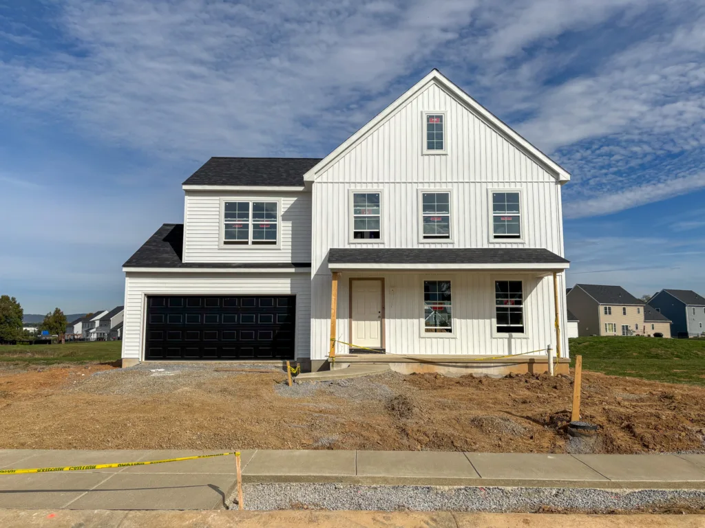 A new two-story white house with black garage doors is under construction in Spring Meadow Reserve, with a dirt yard, gravel, and caution tape in front. Other houses are visible nearby under a blue sky with clouds.