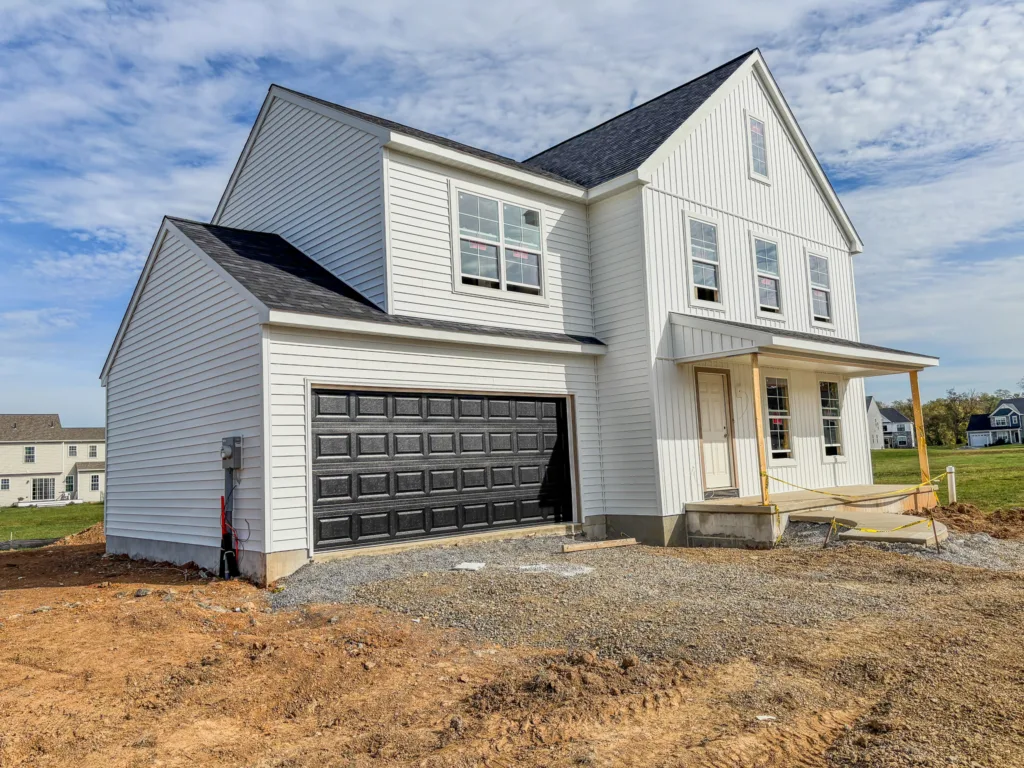 A newly constructed Edison Classic two-story white house with black garage doors and a covered front porch, surrounded by bare soil and gravel, sits in Spring Meadow Reserve with neighboring houses and a blue sky in the background.