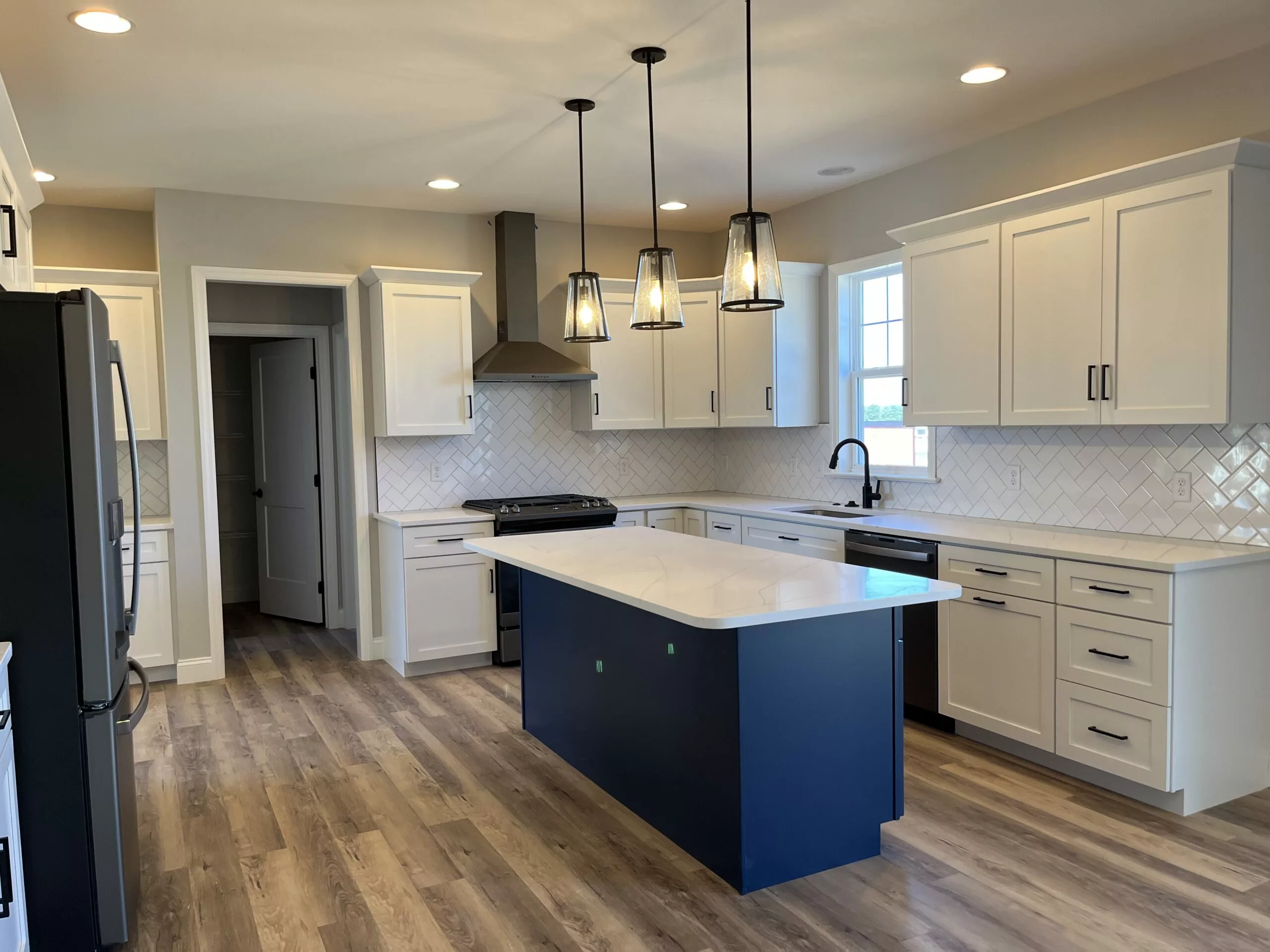 Modern kitchen with white cabinets, stainless steel appliances, a navy blue island with a white countertop, pendant lights above, white tile backsplash, and light wood flooring. Natural light streams in from a window.