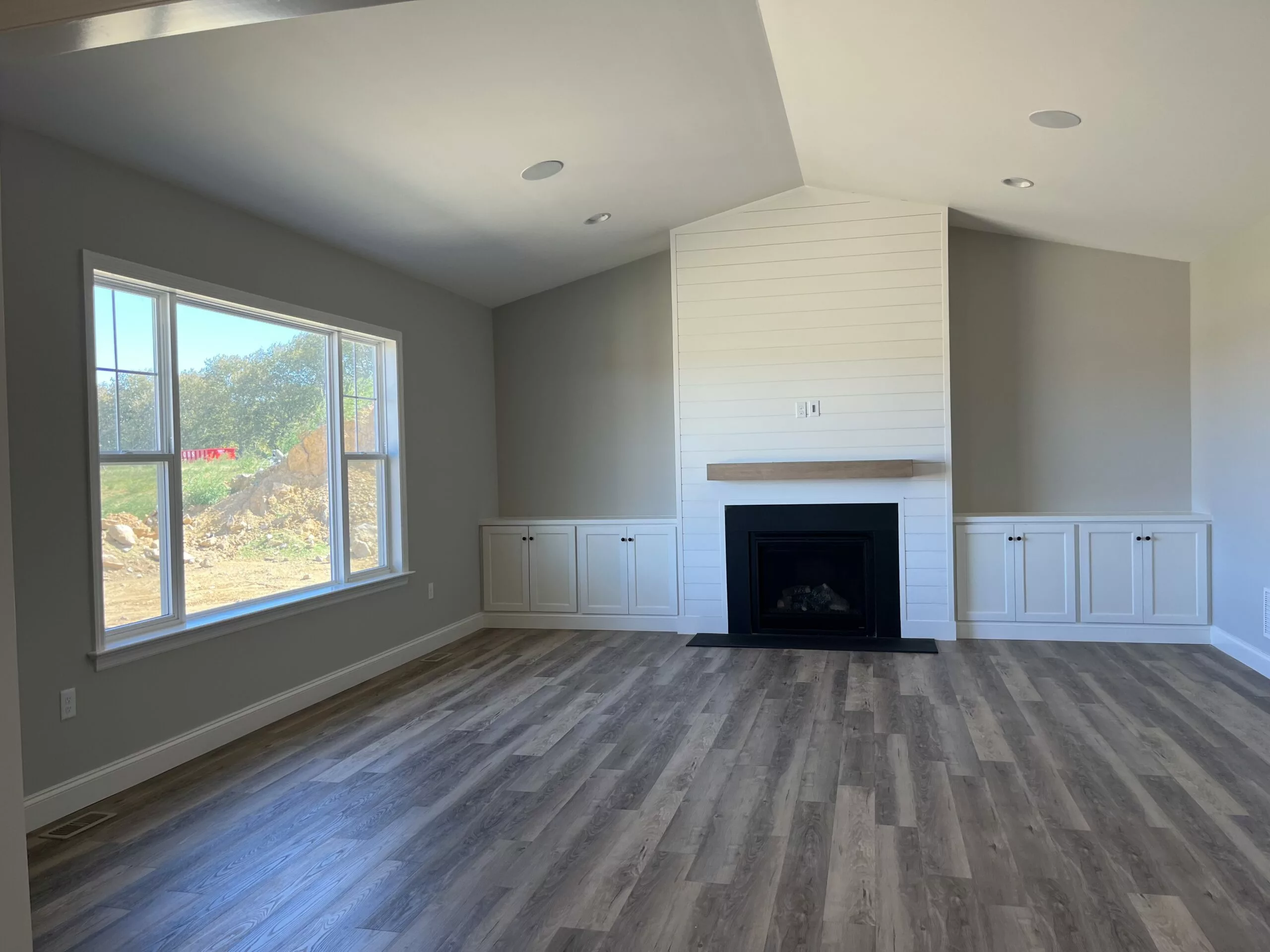 A bright, empty living room with light wood floors, a large window, a white shiplap fireplace with a wooden mantel, and built-in cabinets on both sides. Neutral gray walls and recessed lighting complete the look.