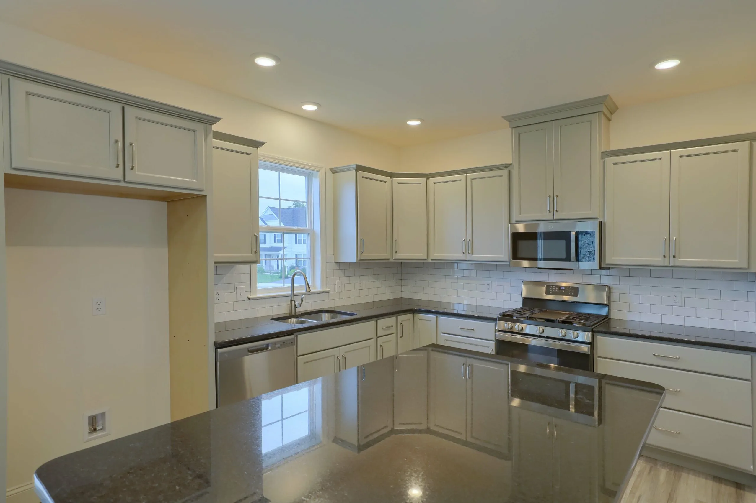 Modern kitchen with light gray cabinets, stainless steel appliances, a gas stove, microwave, dishwasher, white subway tile backsplash, and a large dark granite island under recessed lighting. There is a window above the sink.