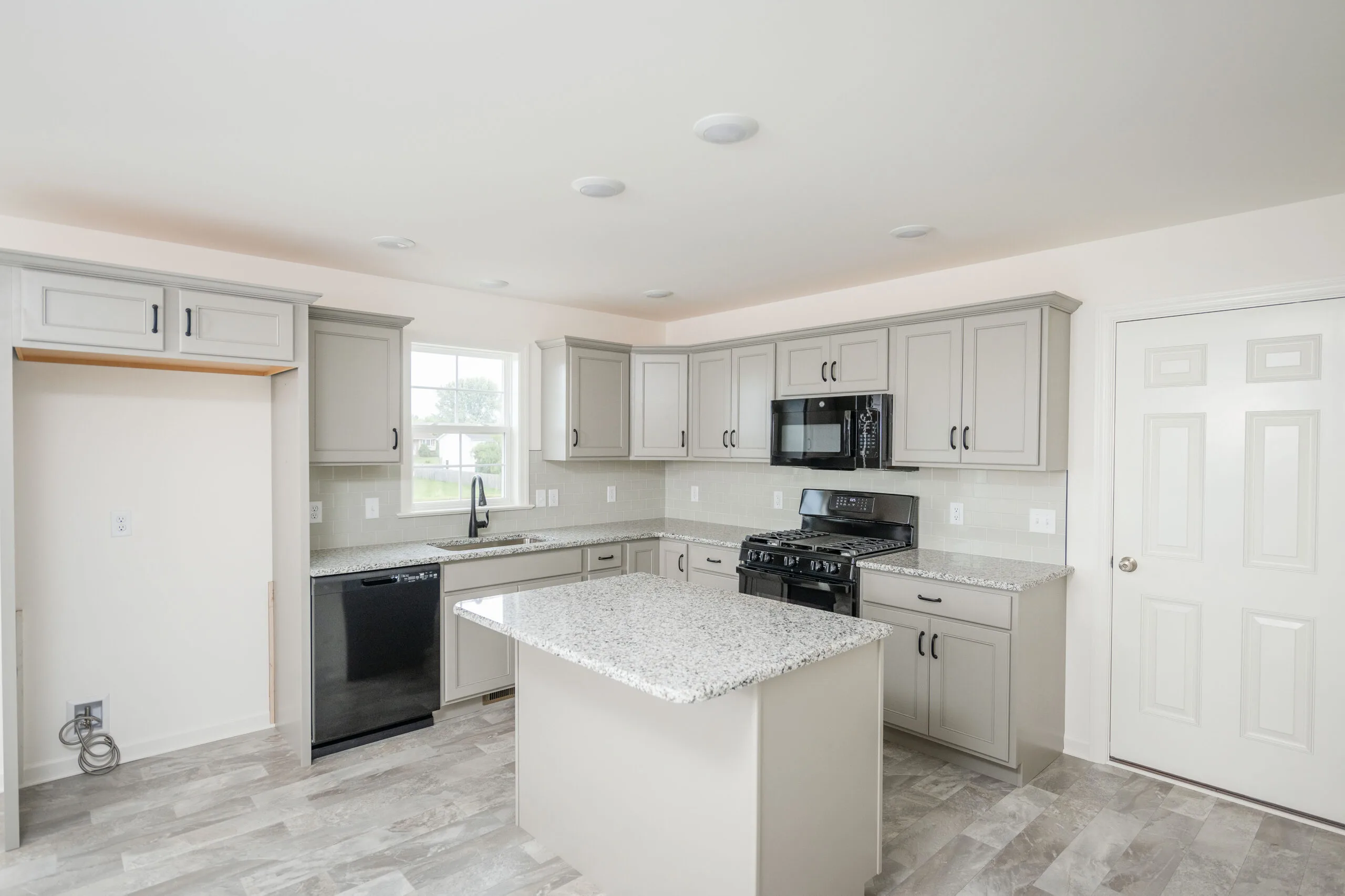 Modern kitchen with light gray cabinets, a granite island, black appliances, a stainless steel sink by a window, and a door to the right. The space is bright with neutral walls and tile flooring.