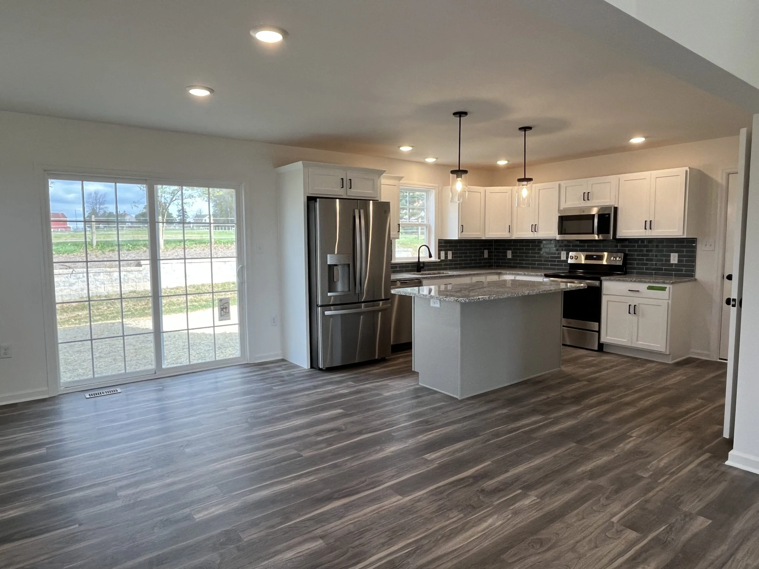 Modern kitchen with white cabinets, stainless steel appliances, a gray island, pendant lighting, dark tile backsplash, and wood-style flooring. Large sliding glass doors open to a patio, letting in natural light.