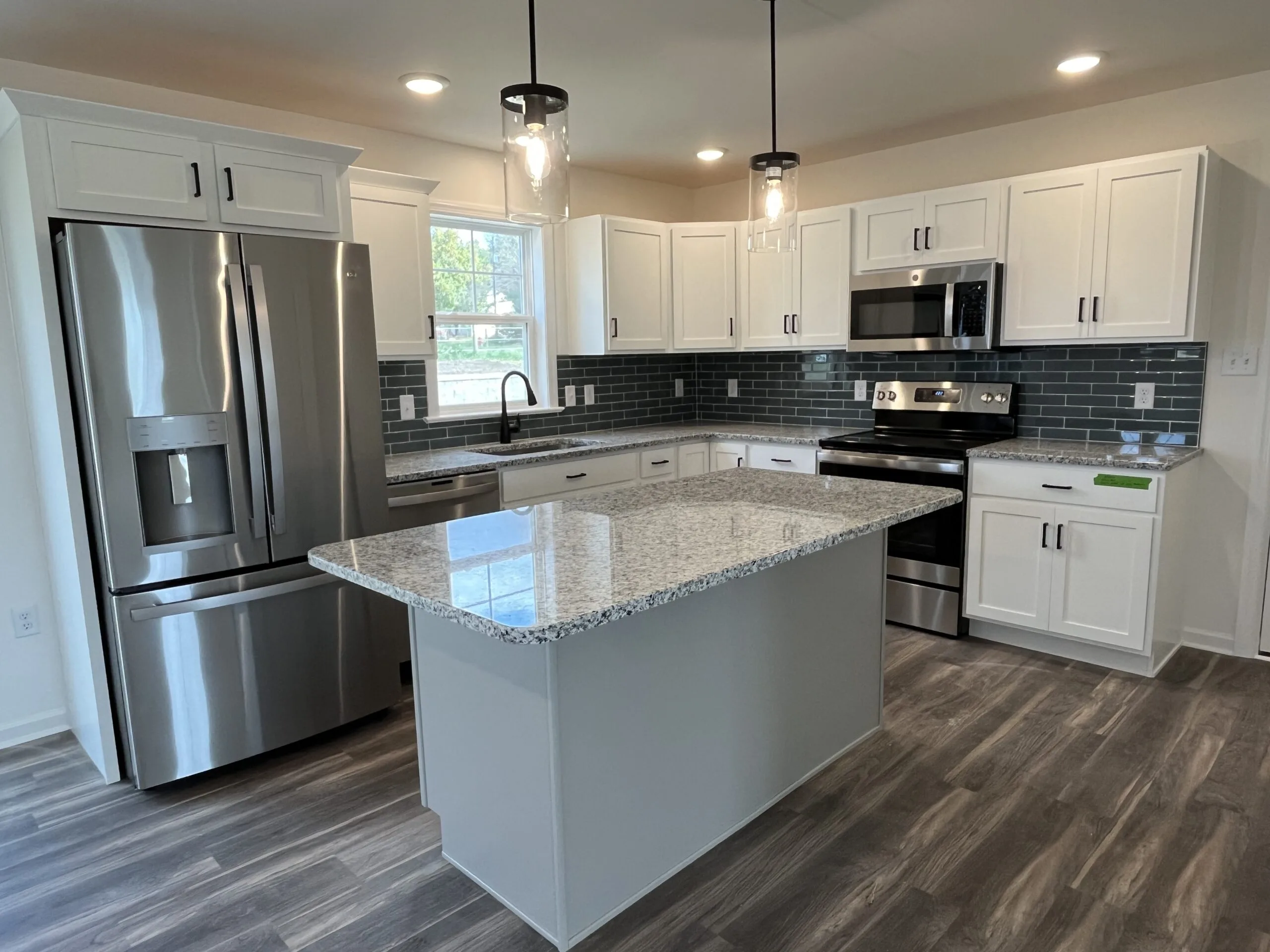 Modern kitchen with white cabinets, a gray tile backsplash, stainless steel appliances, granite countertops, a large island, and pendant lighting. The floor is wood-patterned, and there is a window above the sink.