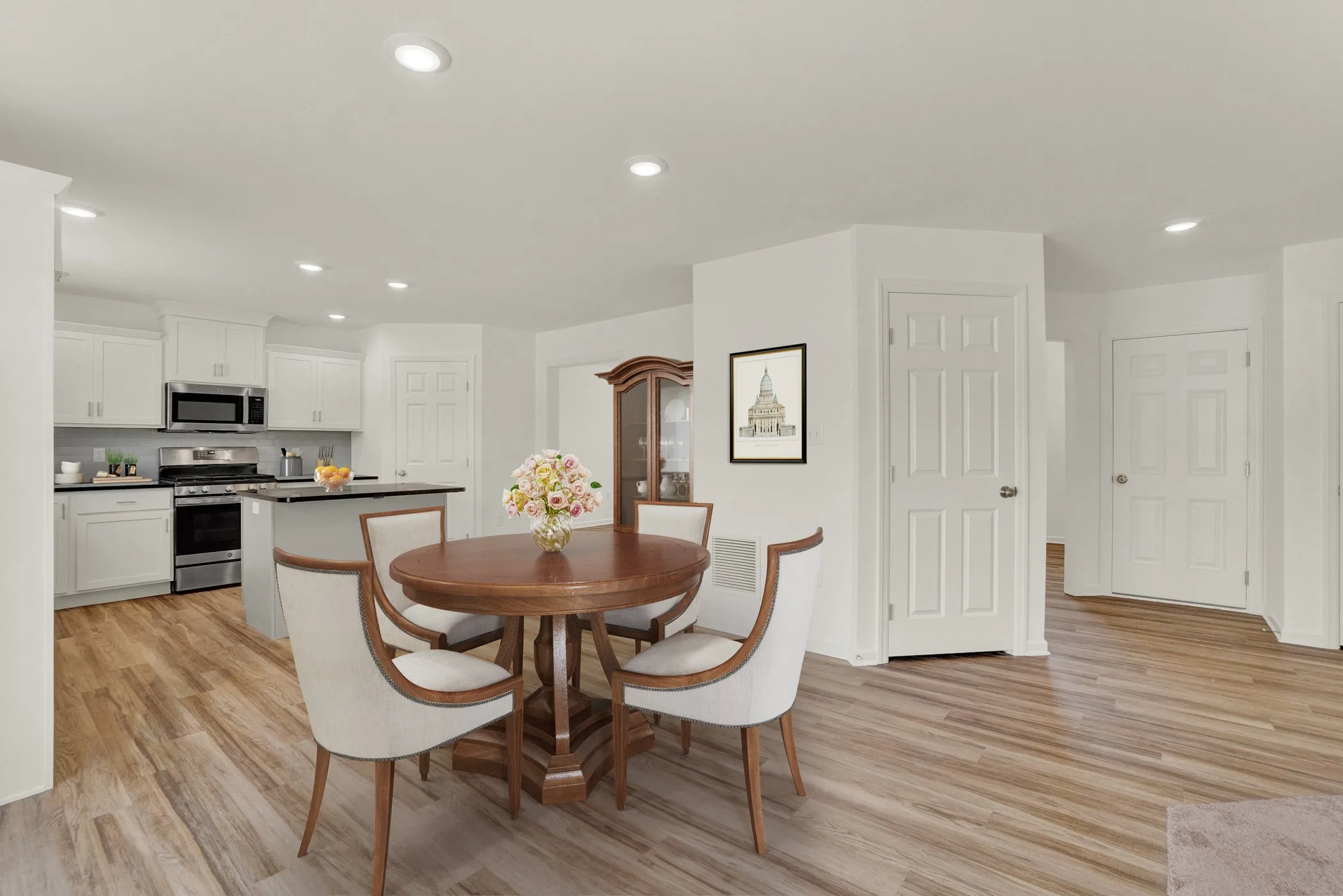 A modern dining area with a round wooden table, four white chairs, a flower centerpiece, and a kitchen with white cabinets and stainless steel appliances in the background. The floor is wood, and walls are white.