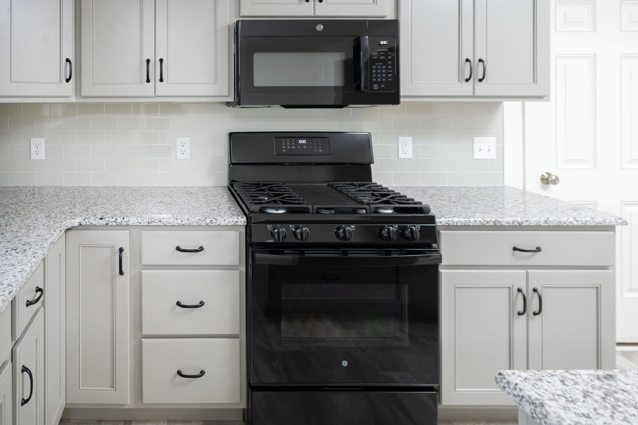 A modern kitchen features a black gas stove and oven with a matching microwave above. The cabinets are light gray with black handles, and the countertops are white with gray speckles. The backsplash is white subway tile.