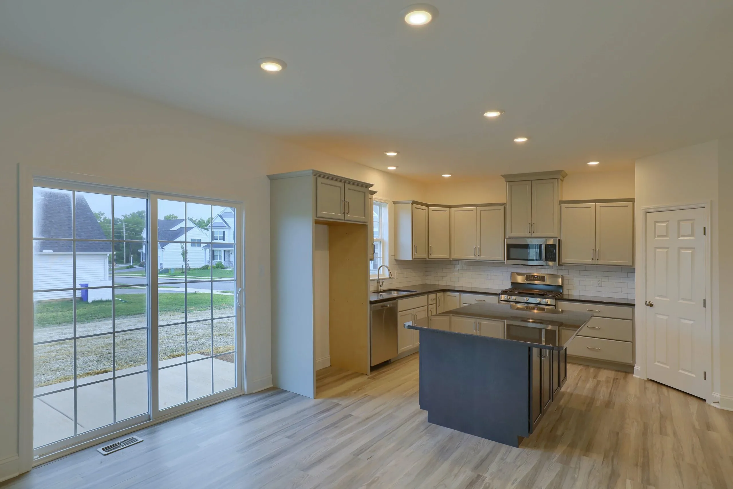 Modern kitchen with light gray cabinets, stainless steel appliances, a central island, wood-style flooring, and recessed lighting. Large sliding glass doors open to a view of a suburban neighborhood outside.