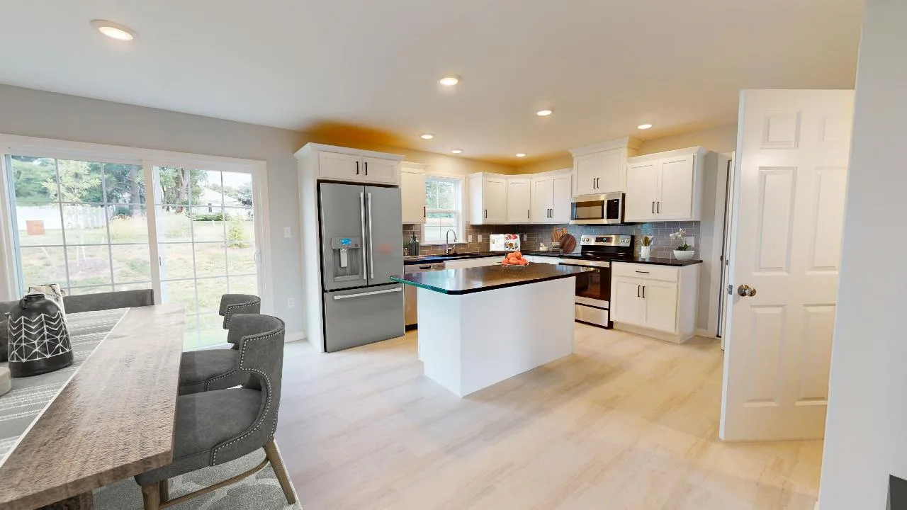 Modern kitchen with white cabinets, stainless steel appliances, a central island with a fruit bowl, dining table with gray chairs, large windows, and light wood flooring. Ample natural light fills the space.