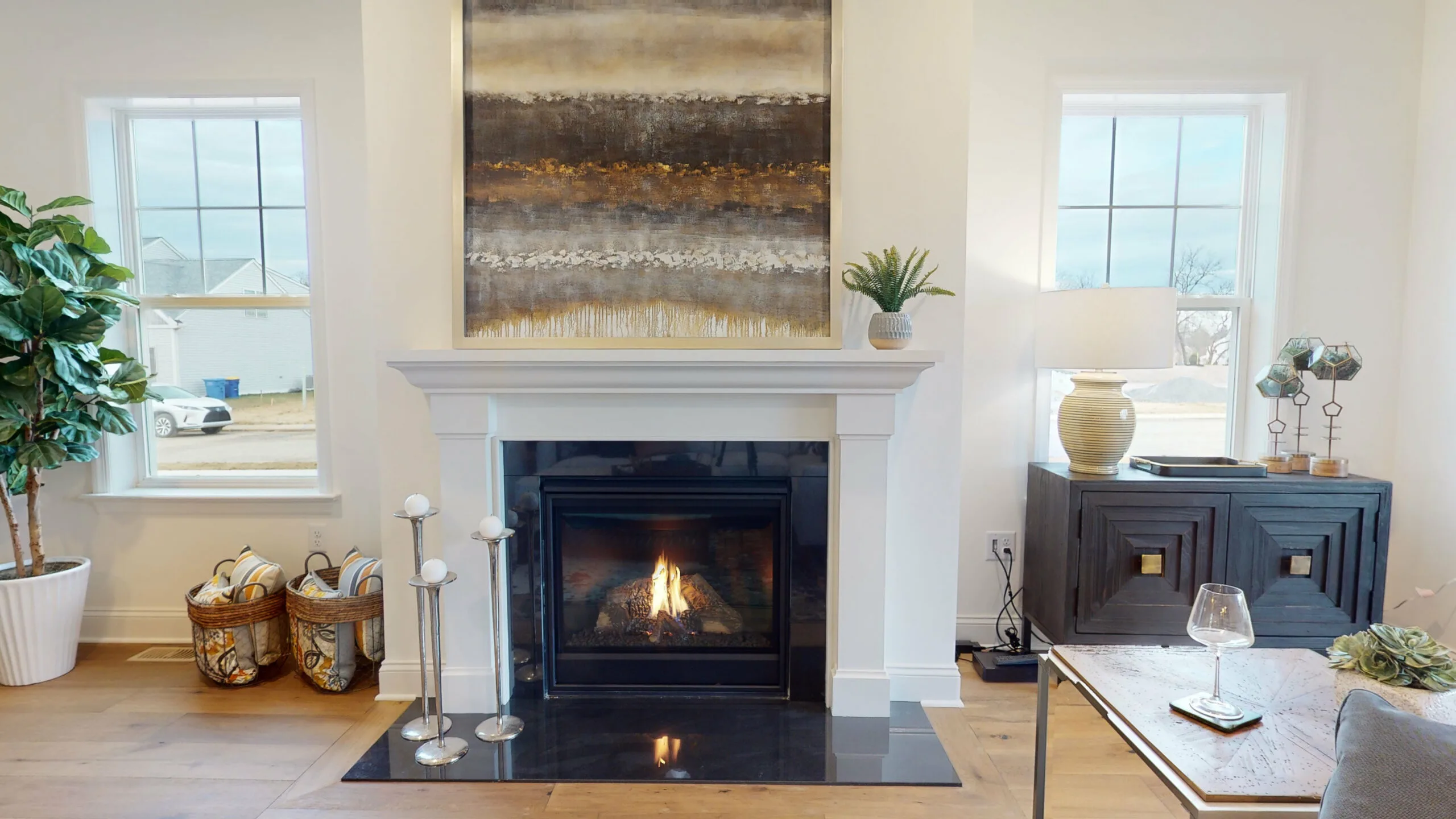 A modern living room with a lit fireplace, abstract artwork above the mantel, and large windows on each side. Decorative plants, woven baskets, and a lamp sit nearby, with a wine glass on a wooden coffee table in front.