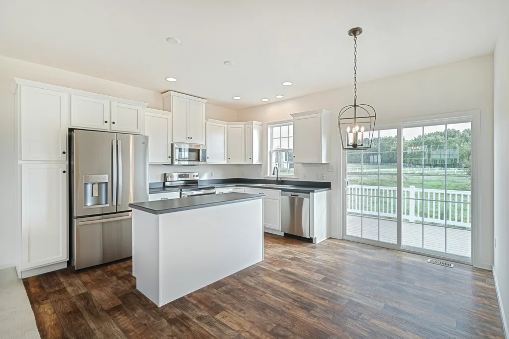 Modern kitchen with white cabinets, stainless steel appliances, a central island, dark countertops, and wood flooring. Large sliding glass doors open to a patio with a view of green grass and trees outside.
