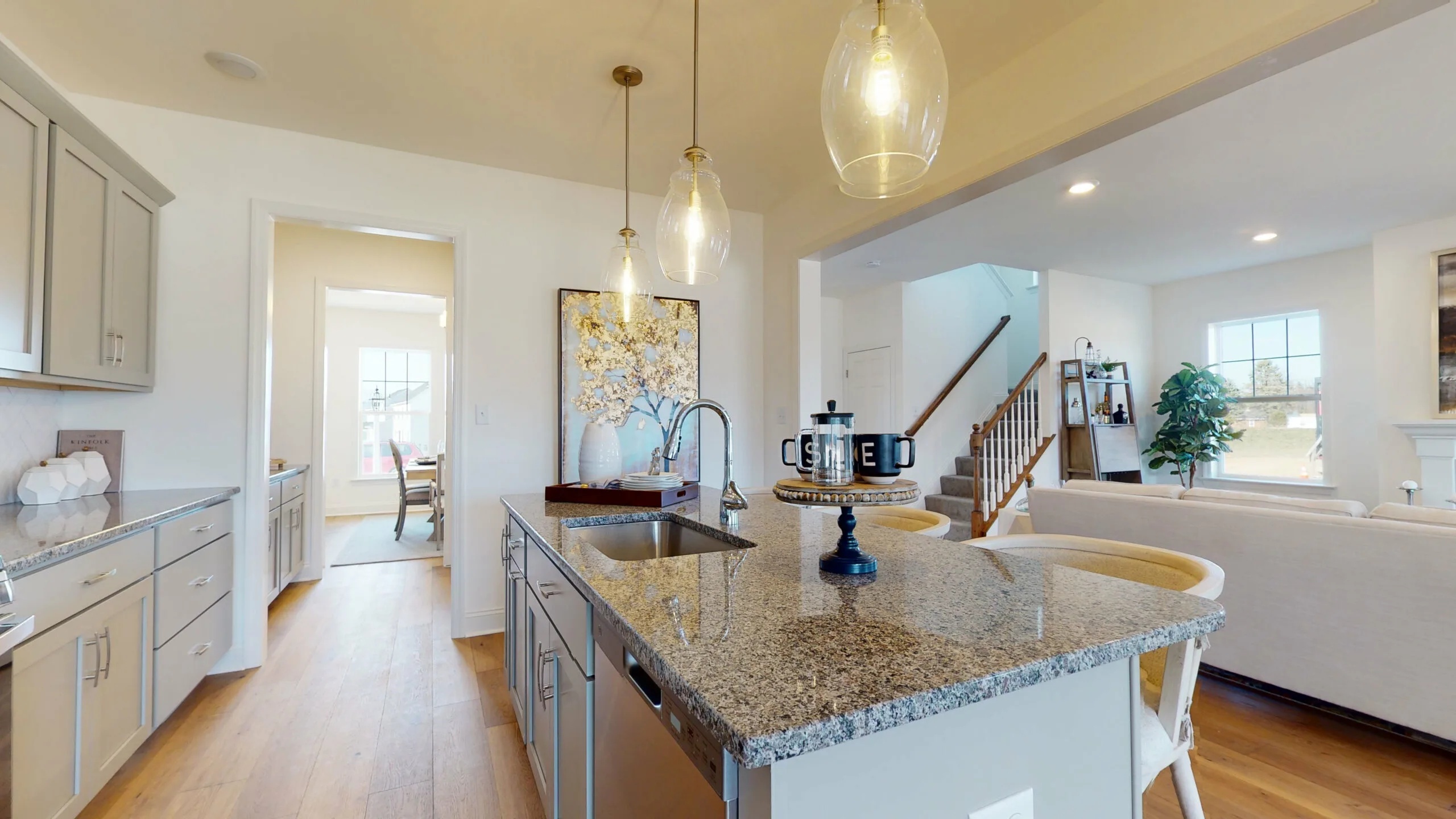 Modern kitchen with granite island, pendant lights, stainless steel sink, and dishwasher. Open layout leads to a bright dining area and living room with staircase and decorative shelving in the background.