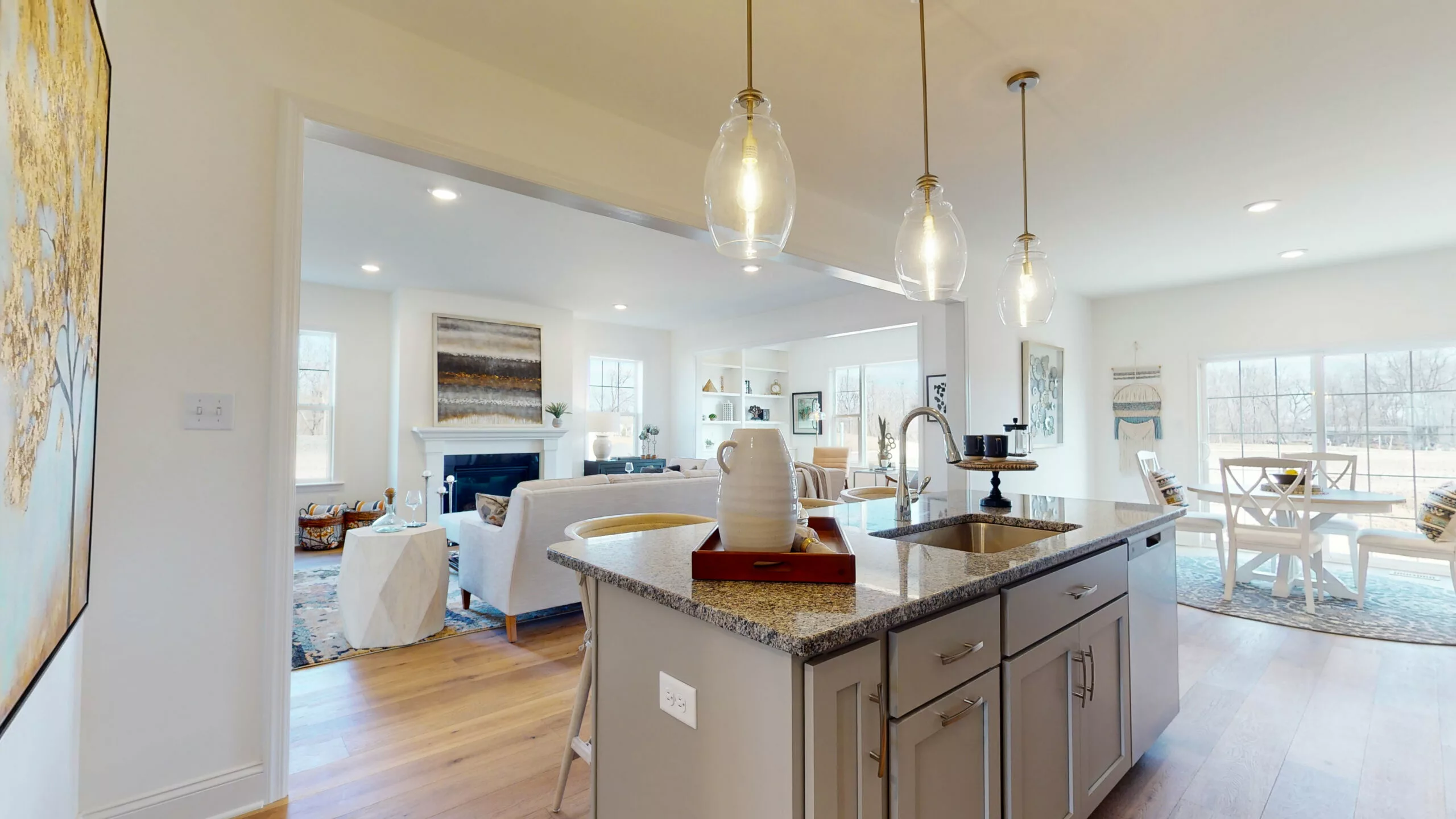 Modern open-concept kitchen and living area with pendant lights, granite island, light gray cabinets, and cozy seating. Large windows let in natural light, with dining table visible in the background.