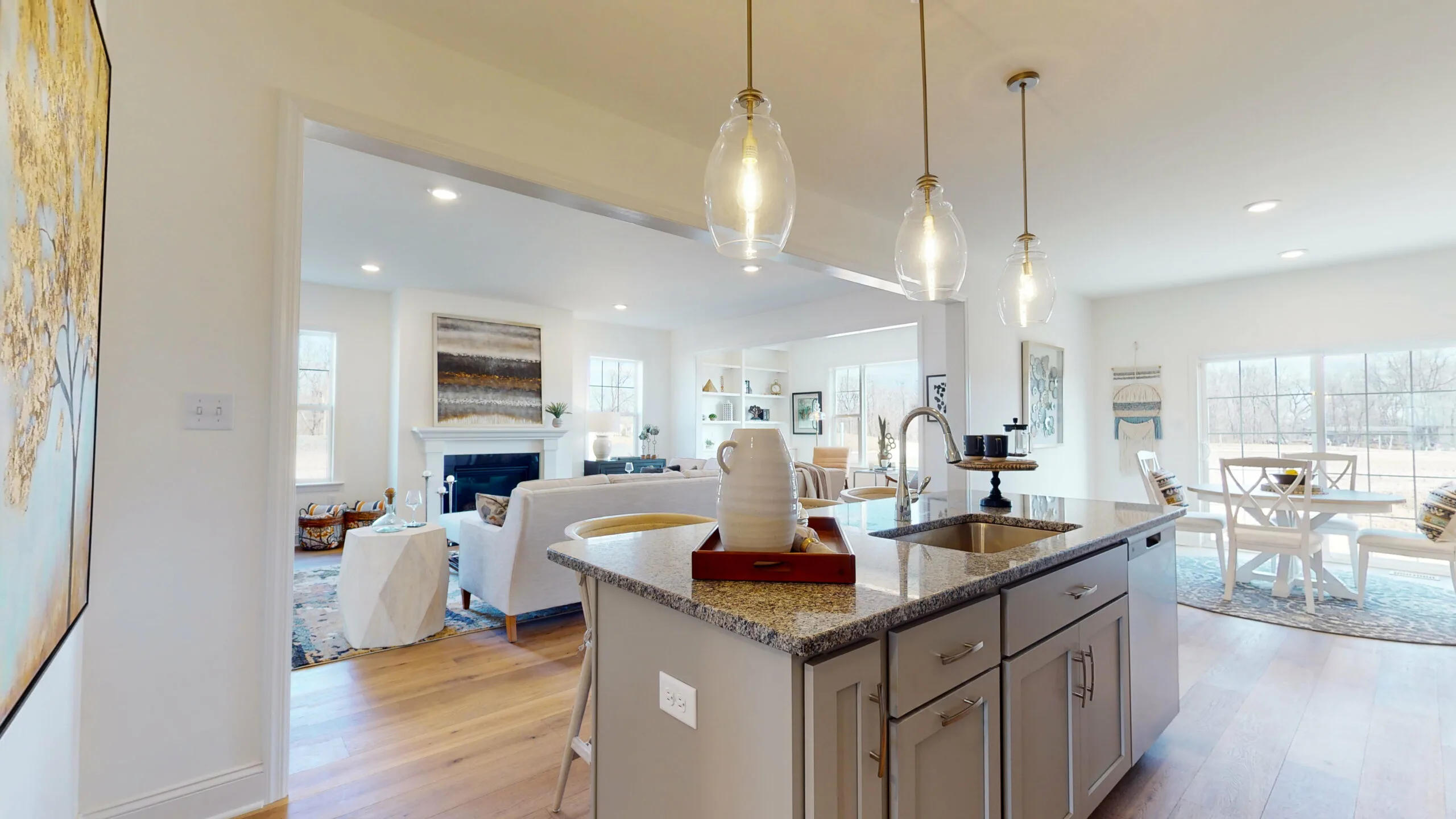 Bright, modern open-concept kitchen with granite island, pendant lights, and sink, overlooking a cozy living room with sofas, fireplace, and large windows, plus a dining area in the background.