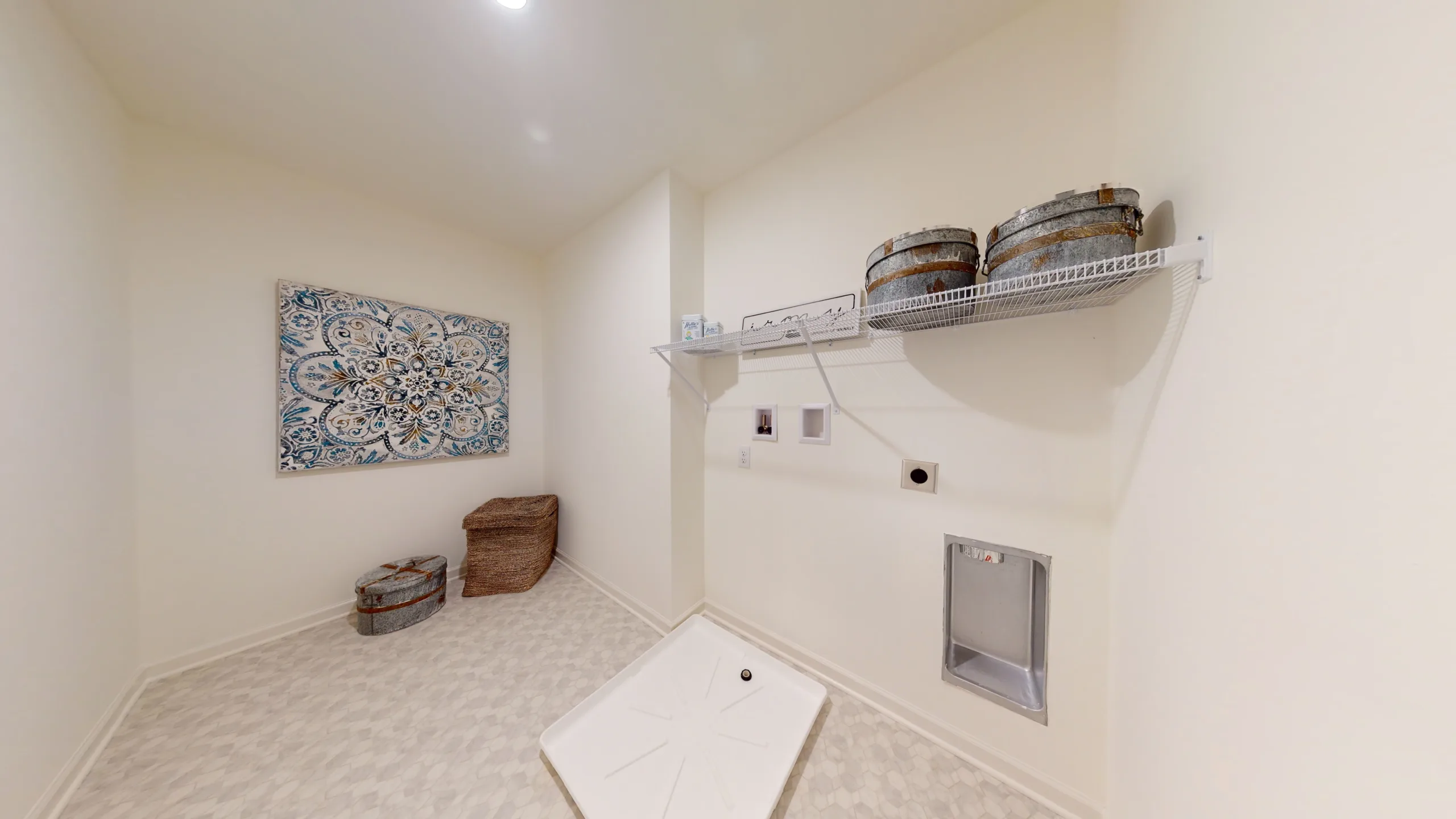 A laundry room with light walls and patterned floor, featuring a washer/dryer hookup, wire shelf with metal baskets, wicker baskets on the floor, a decorative wall art, and a small recessed utility box.
