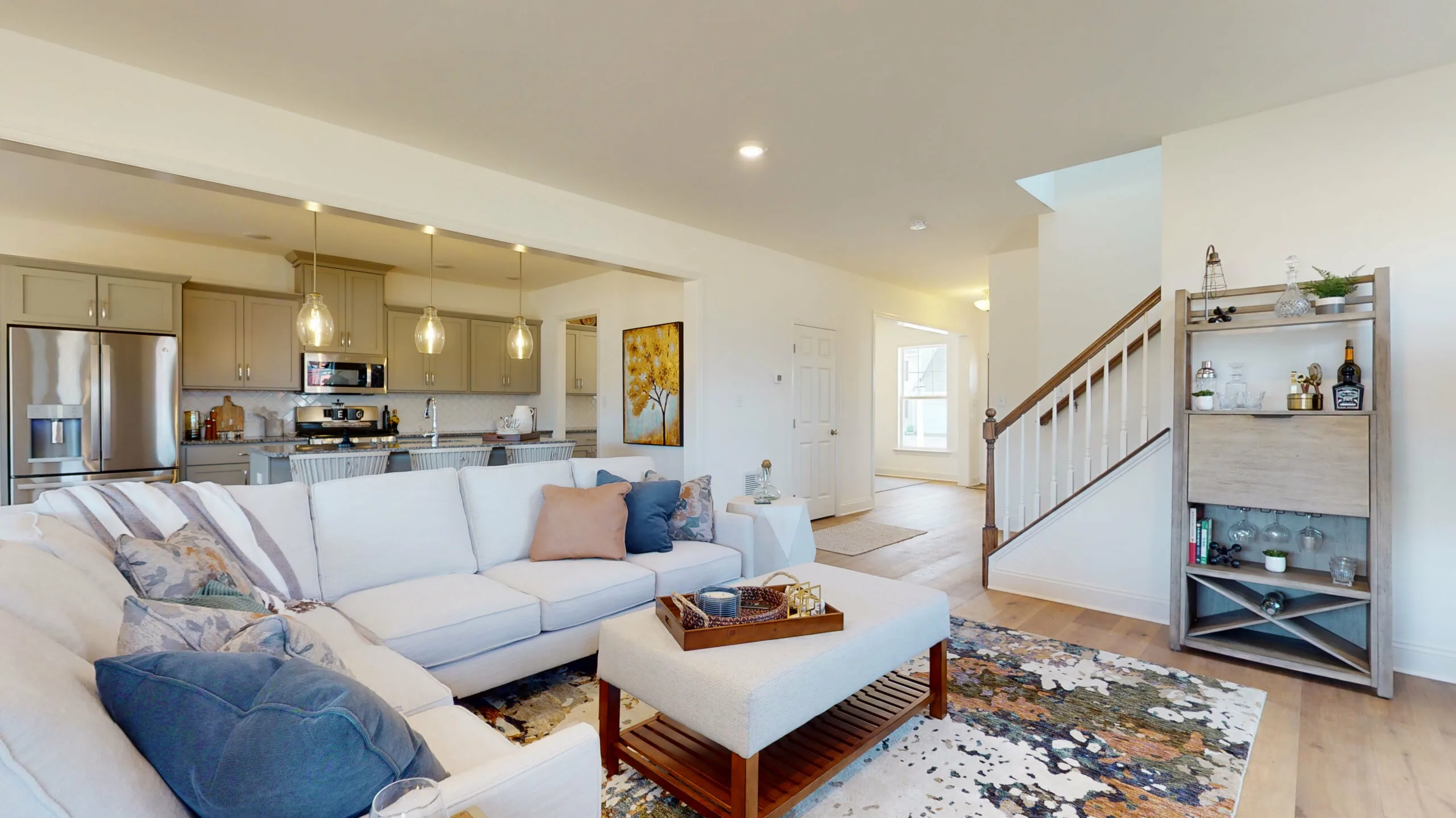 Modern open-concept living room with a white sectional sofa, blue and brown accents, ottoman, and colorful rug. Kitchen with stainless steel appliances in the background, stairs and bookshelf to the right. Bright and airy atmosphere.