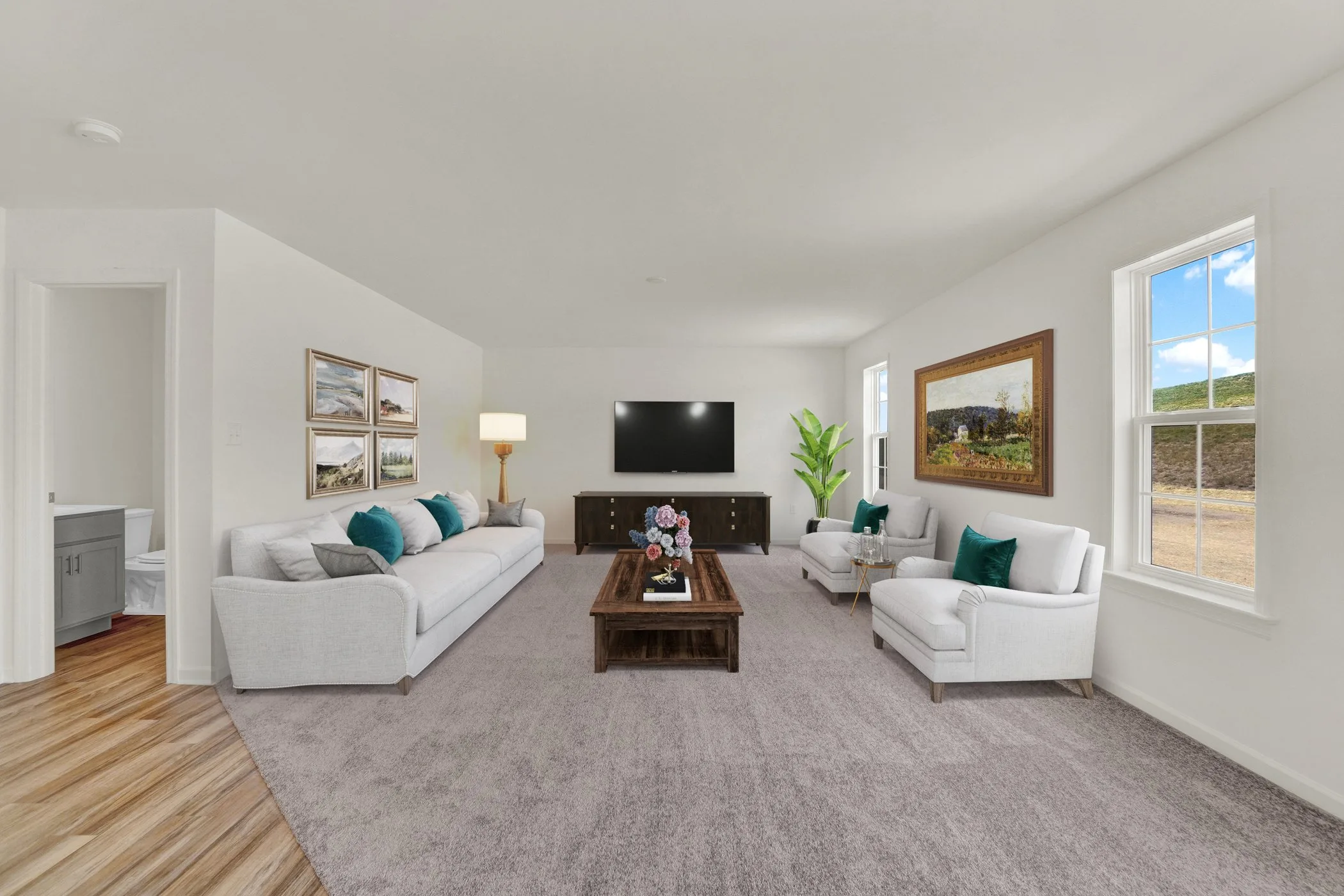 A spacious, bright living room with white walls, large windows, two white sofas, two armchairs, a wooden coffee table, a TV, and wall art. The room has light wood and carpeted flooring and a plant in the corner.