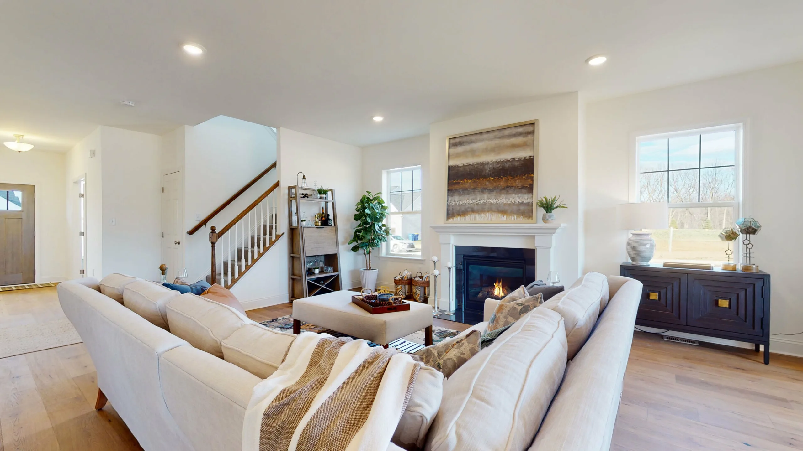 Bright living room with a beige sectional sofa, striped throw, coffee table, fireplace with art above, large windows, bookshelf, potted plant, and staircase in the background. Natural light fills the space.