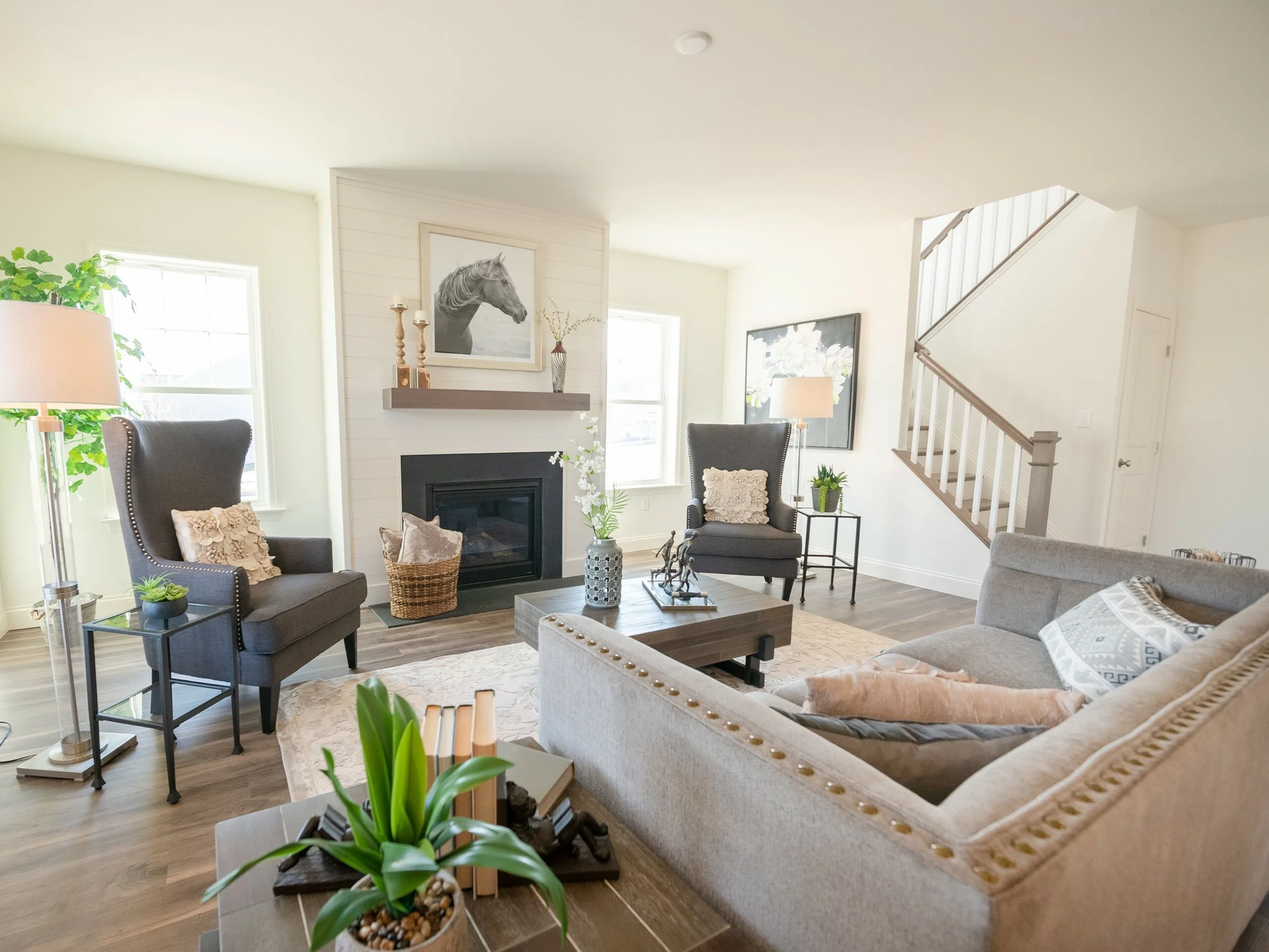 Bright, modern living room with a gray sofa and two armchairs, a fireplace with a horse artwork above it, large windows, potted plants, and a staircase in the background. Neutral decor and wood flooring.