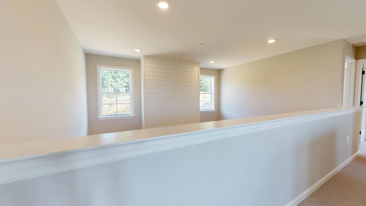 A bright upstairs hallway with beige walls and carpet, white trim, recessed ceiling lights, and two windows at the end letting in natural light.