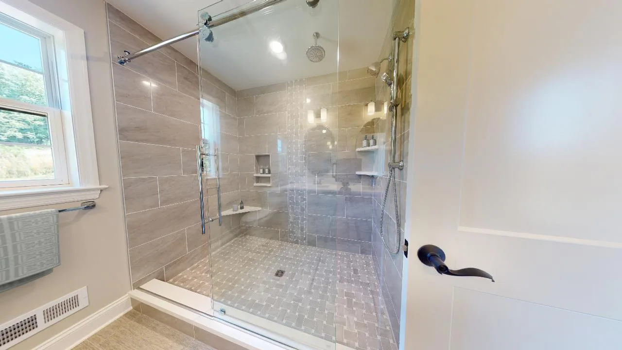 A spacious bathroom with a large glass-enclosed walk-in shower, beige tile walls, a rainfall showerhead, built-in corner shelves with toiletries, and natural light coming in from a window.
