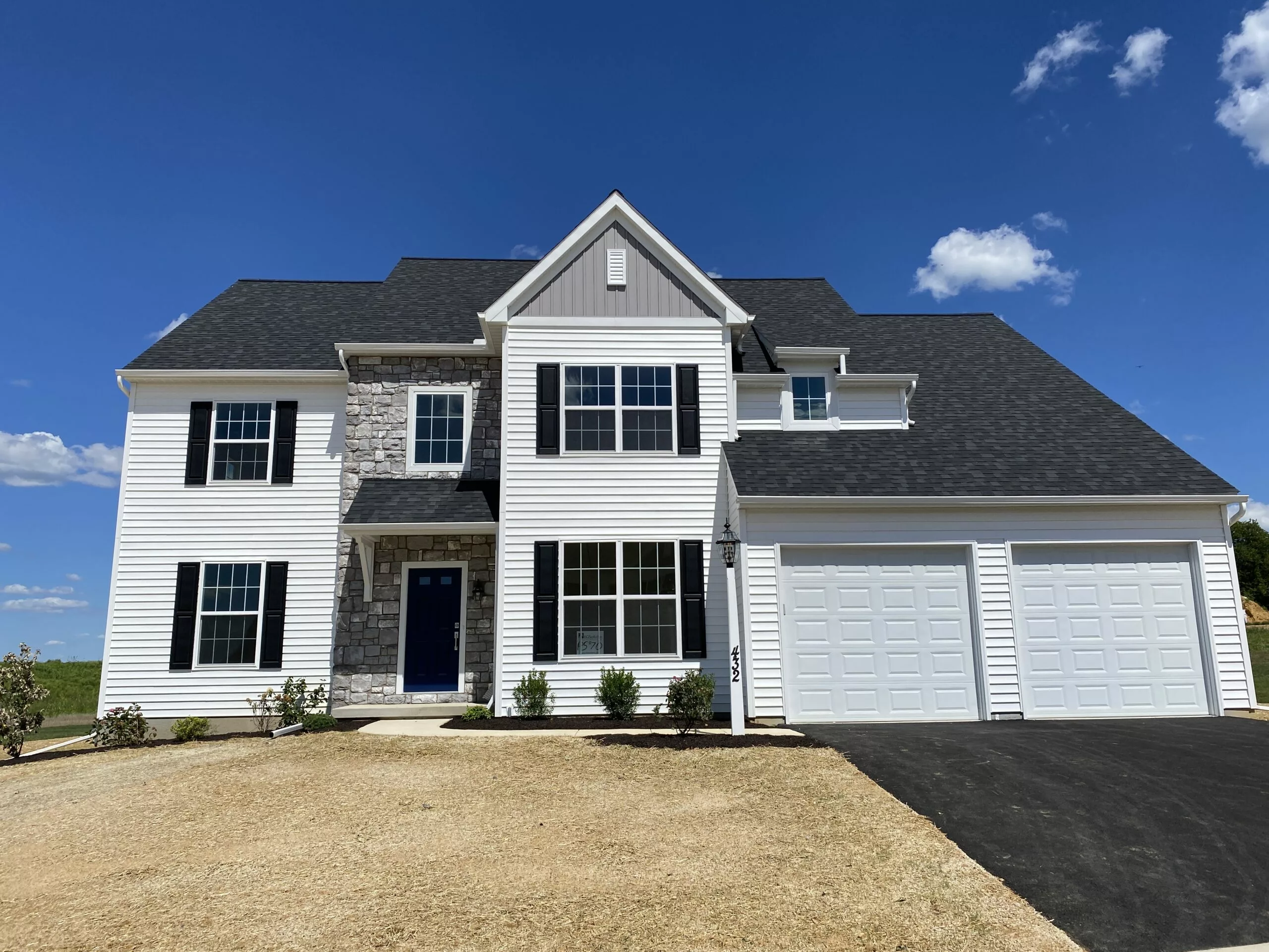 A two-story suburban house with white siding, stone accents, black shutters, and a dark roof. There are two garage doors, a driveway, a blue front door, and a lawn with sparse, dry grass under a clear blue sky.