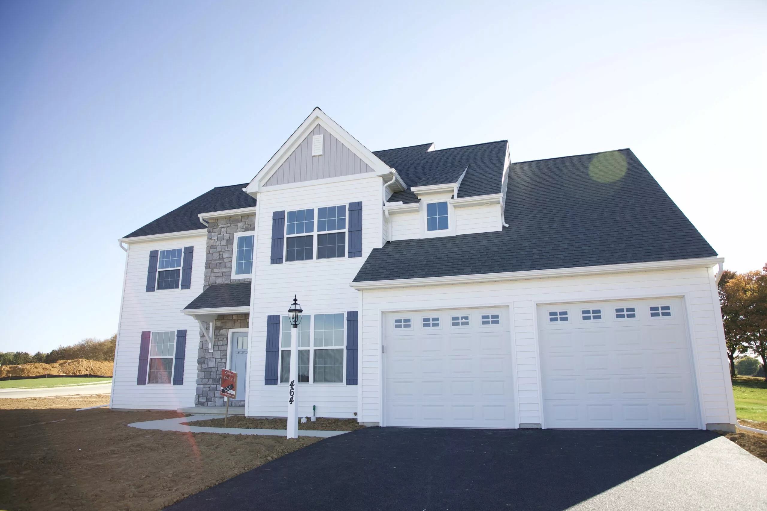 A modern two-story suburban house with white siding, gray stone accents, large windows, and a two-car garage. The driveway is paved, and the yard is freshly landscaped with minimal grass.