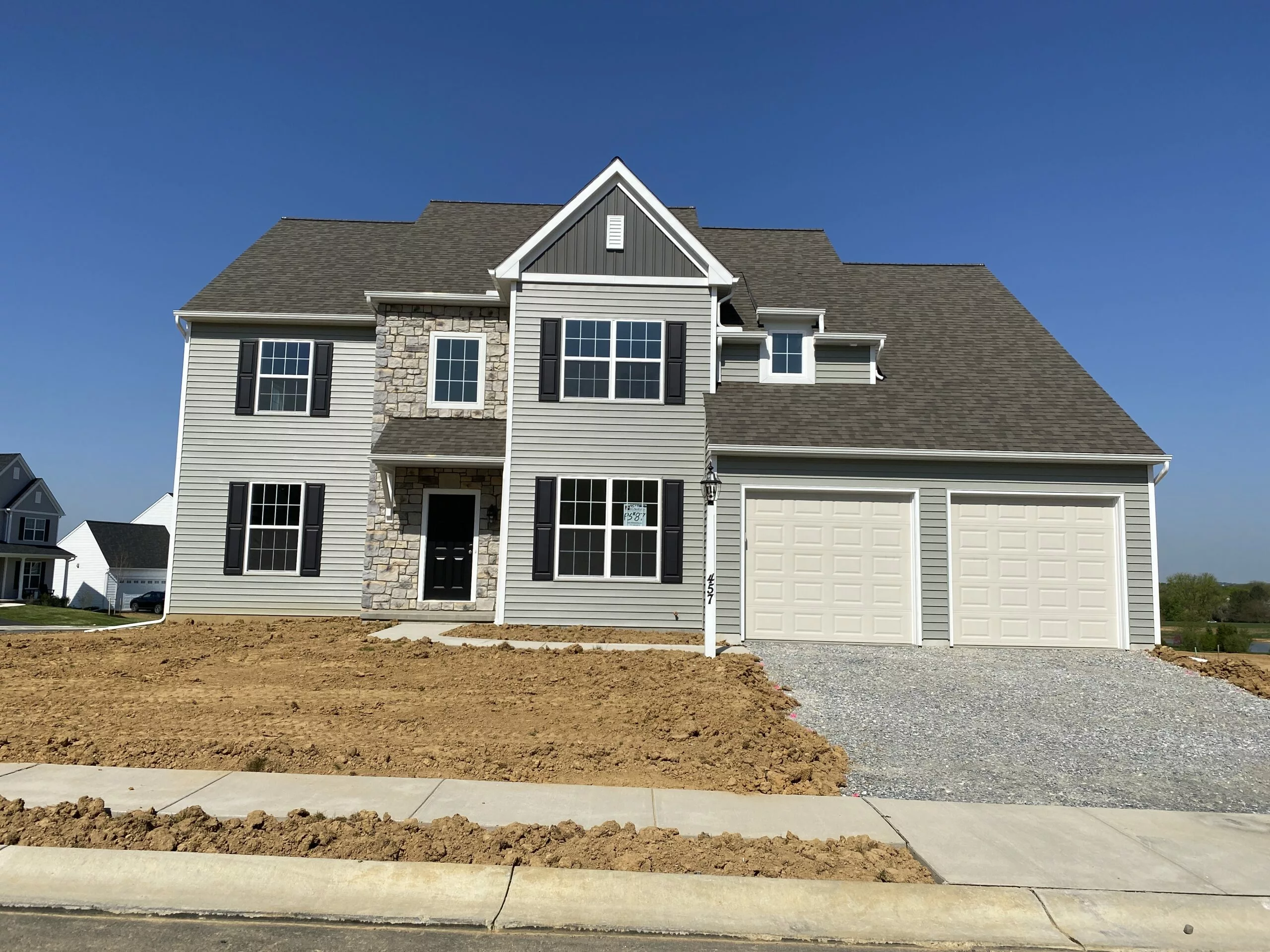 A newly constructed two-story house with gray siding, stone accents, and a large two-car garage. The yard is bare dirt with no landscaping yet, and the sky is clear and blue.