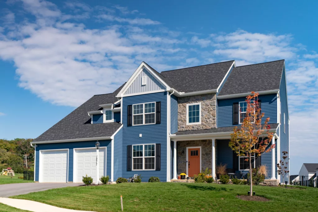 A modern two-story house with blue siding, stone accents, and white trim, featuring a double garage, front porch with an orange door, and a neatly landscaped yard under a partly cloudy sky.