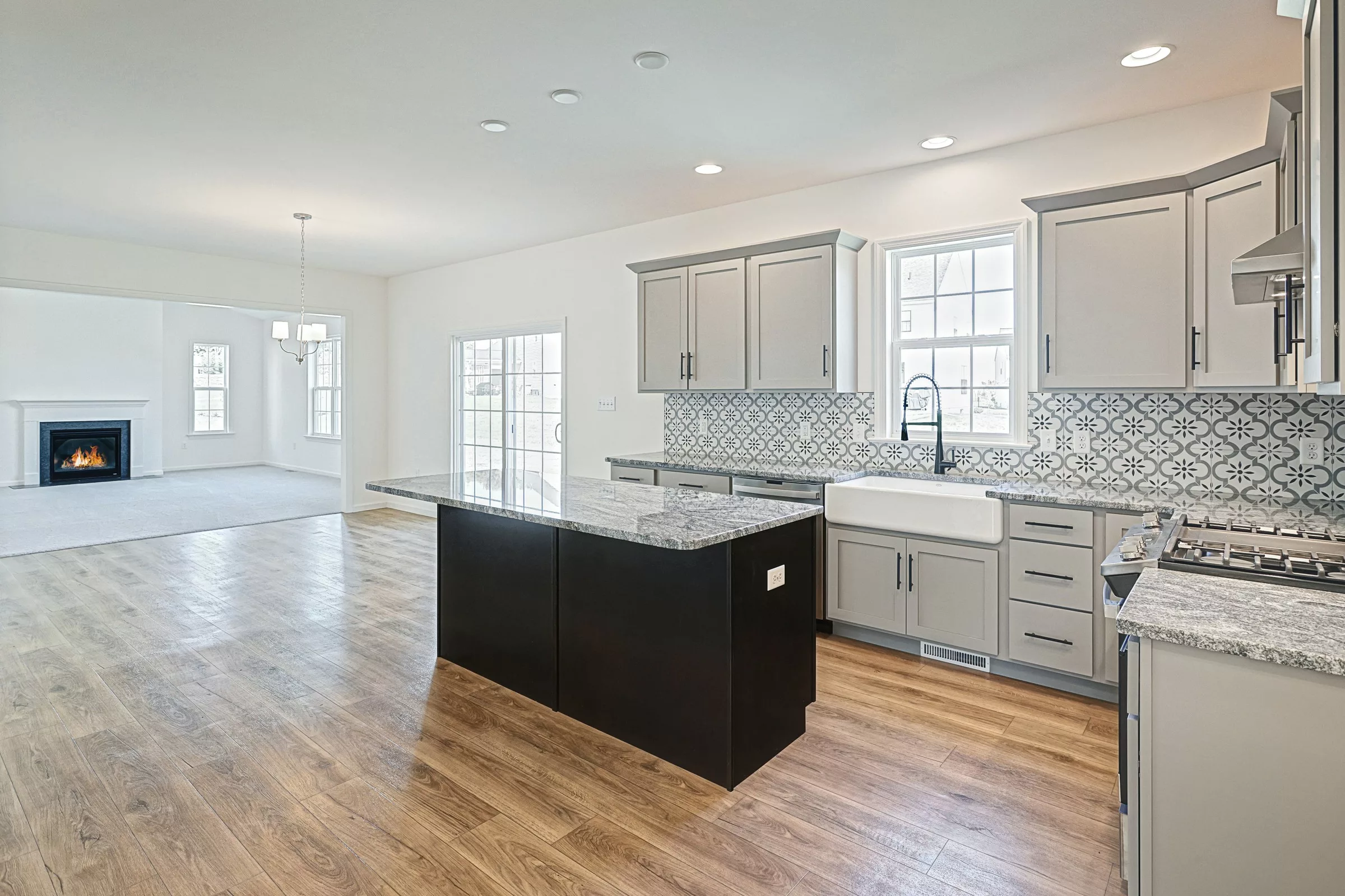 Modern kitchen with gray cabinets, patterned tile backsplash, granite countertops, and a central island. Open floor plan leads to a bright living area with a fireplace and large windows. Light wood floors throughout.