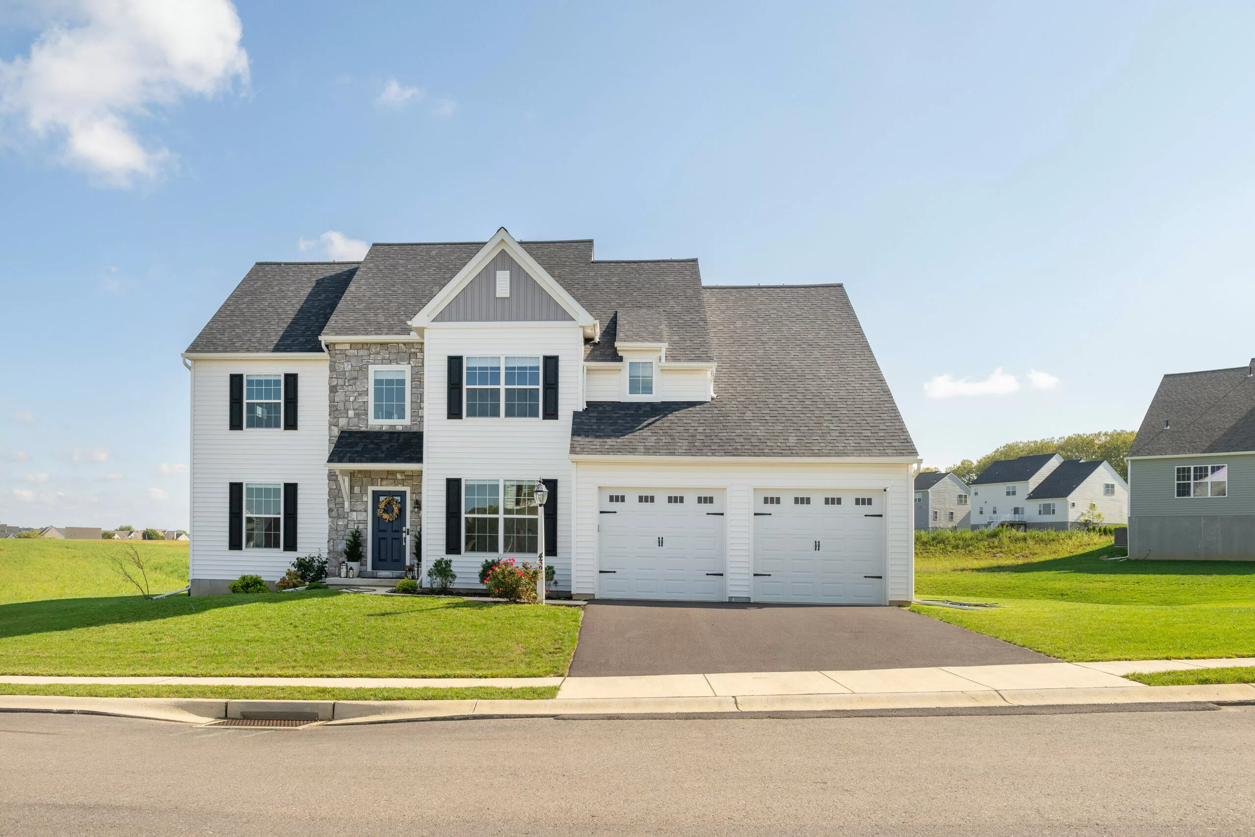 A two-story suburban house with white siding, black shutters, and a stone-accented entrance, featuring a three-car garage and a well-kept lawn under a clear blue sky.