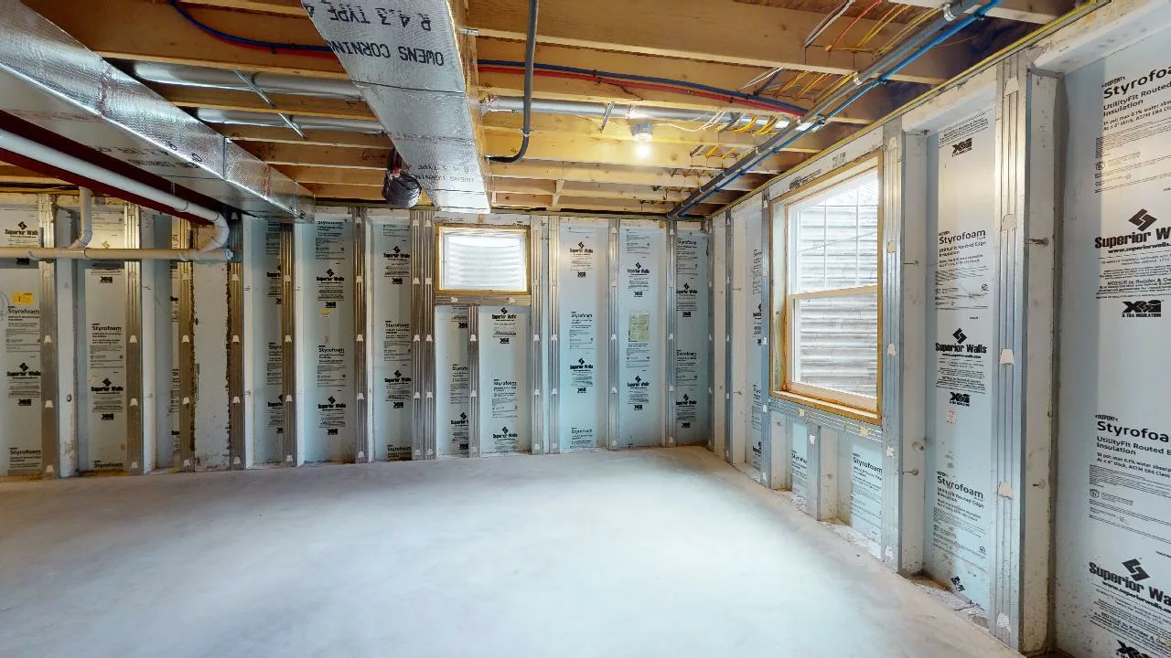 Unfinished basement room with exposed insulation panels on the walls, visible ceiling beams, ductwork, and wiring; light coming through two windows, and a bare concrete floor.
