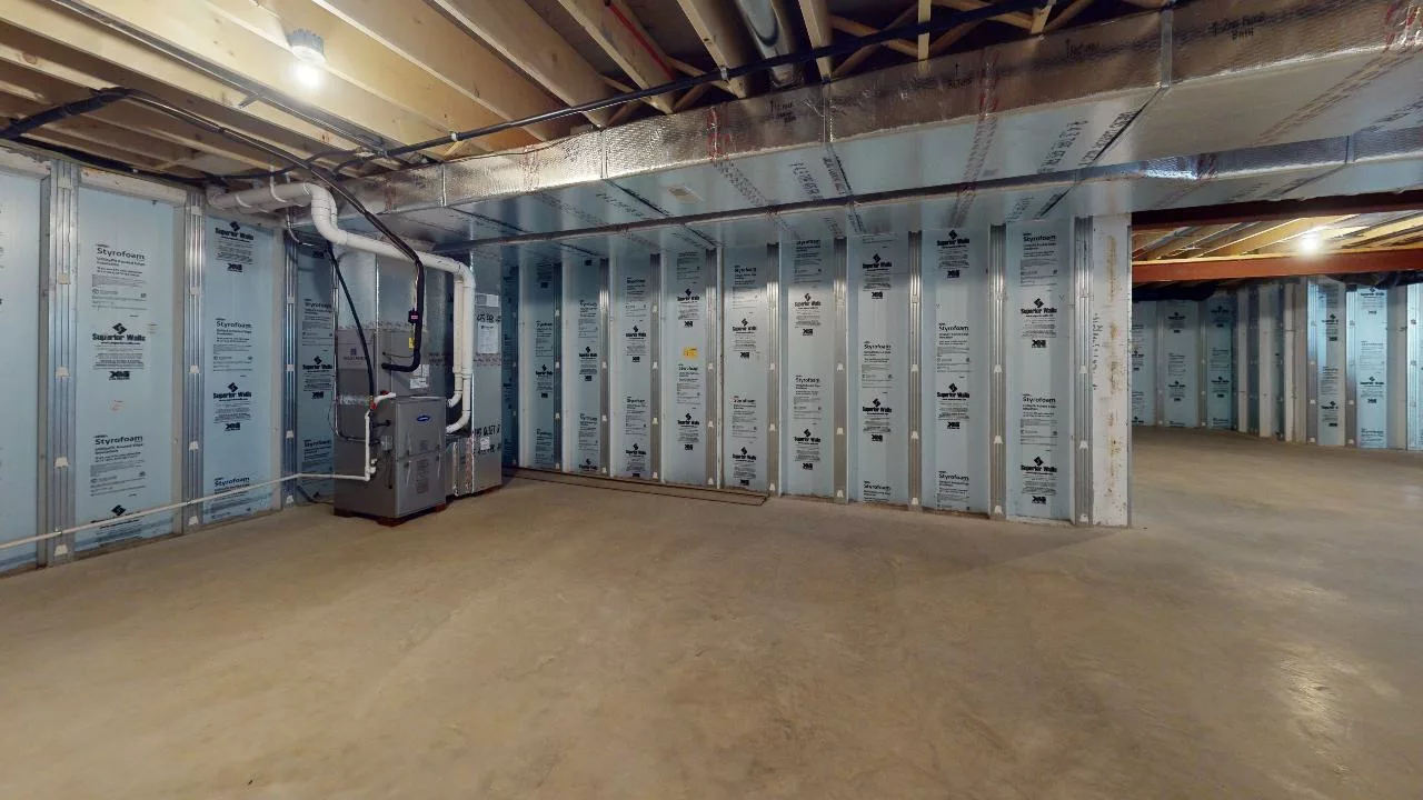 Spacious unfinished basement with exposed ceiling beams, concrete floor, insulated walls with visible insulation panels, and a furnace unit in one corner. Brightly lit by ceiling lights.