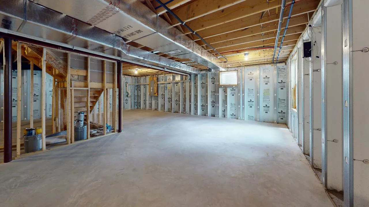 Unfinished basement with concrete floor, exposed wooden ceiling beams, metal ductwork, insulated walls, and wooden stairs leading to the main floor. Natural light enters through a small window.