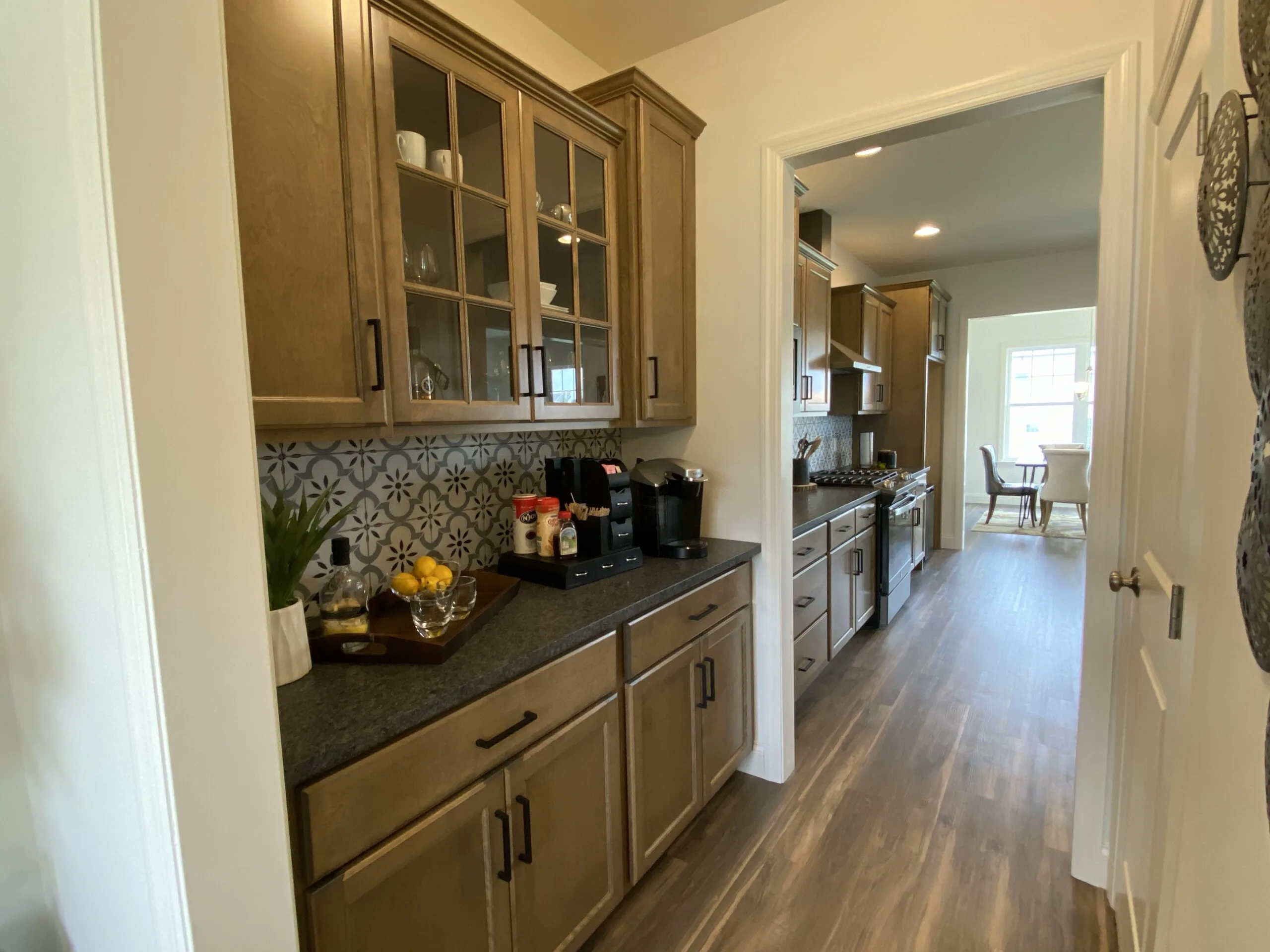 A narrow kitchen with light wood cabinets, glass doors, a black countertop, patterned backsplash, coffee maker, and tray with glasses. The kitchen leads to a dining area with a window in the background.