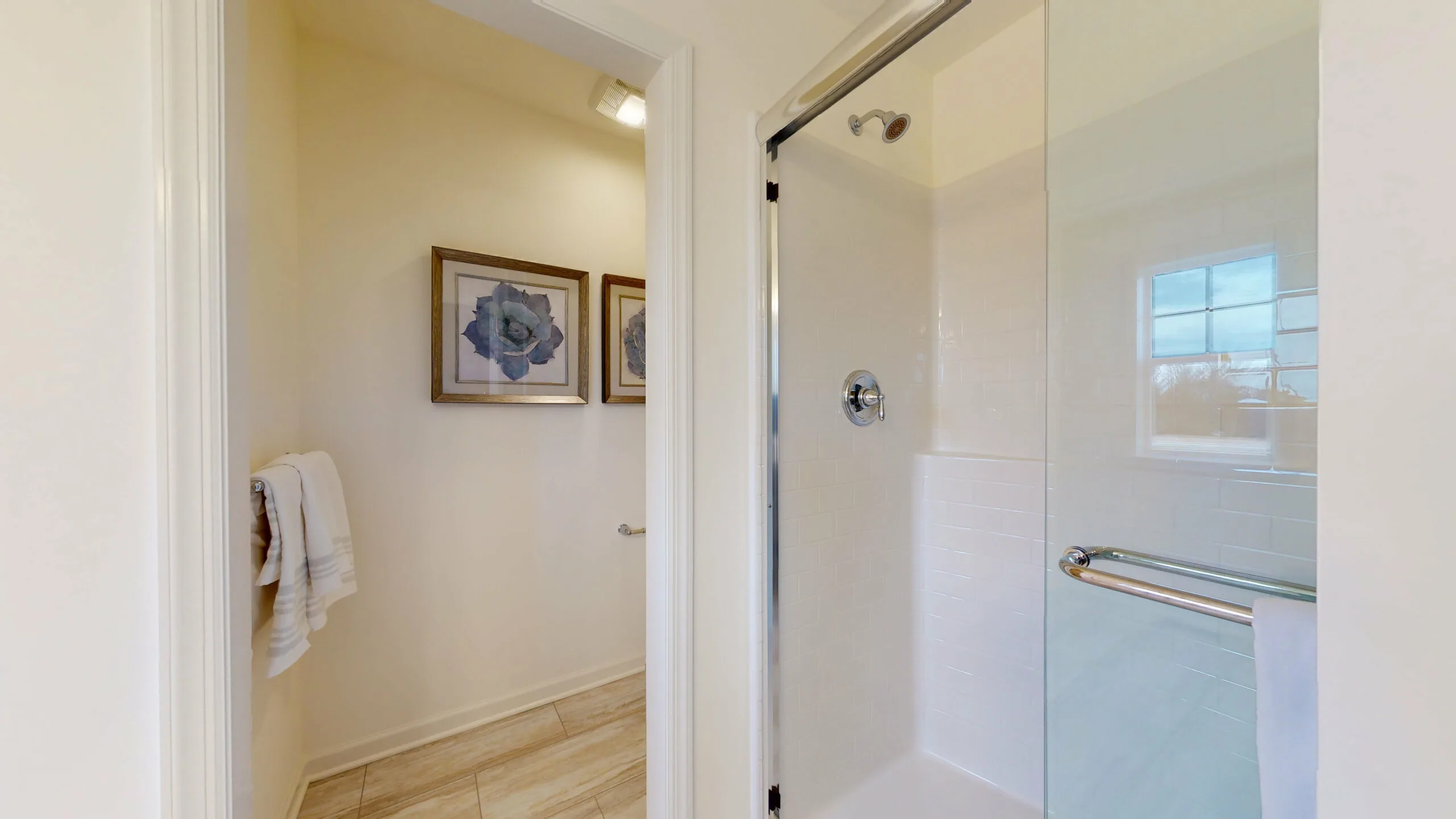 A bright bathroom with a glass shower enclosure, beige tile floor, and two framed floral pictures on the wall. A white towel hangs on a rack beneath the artwork.