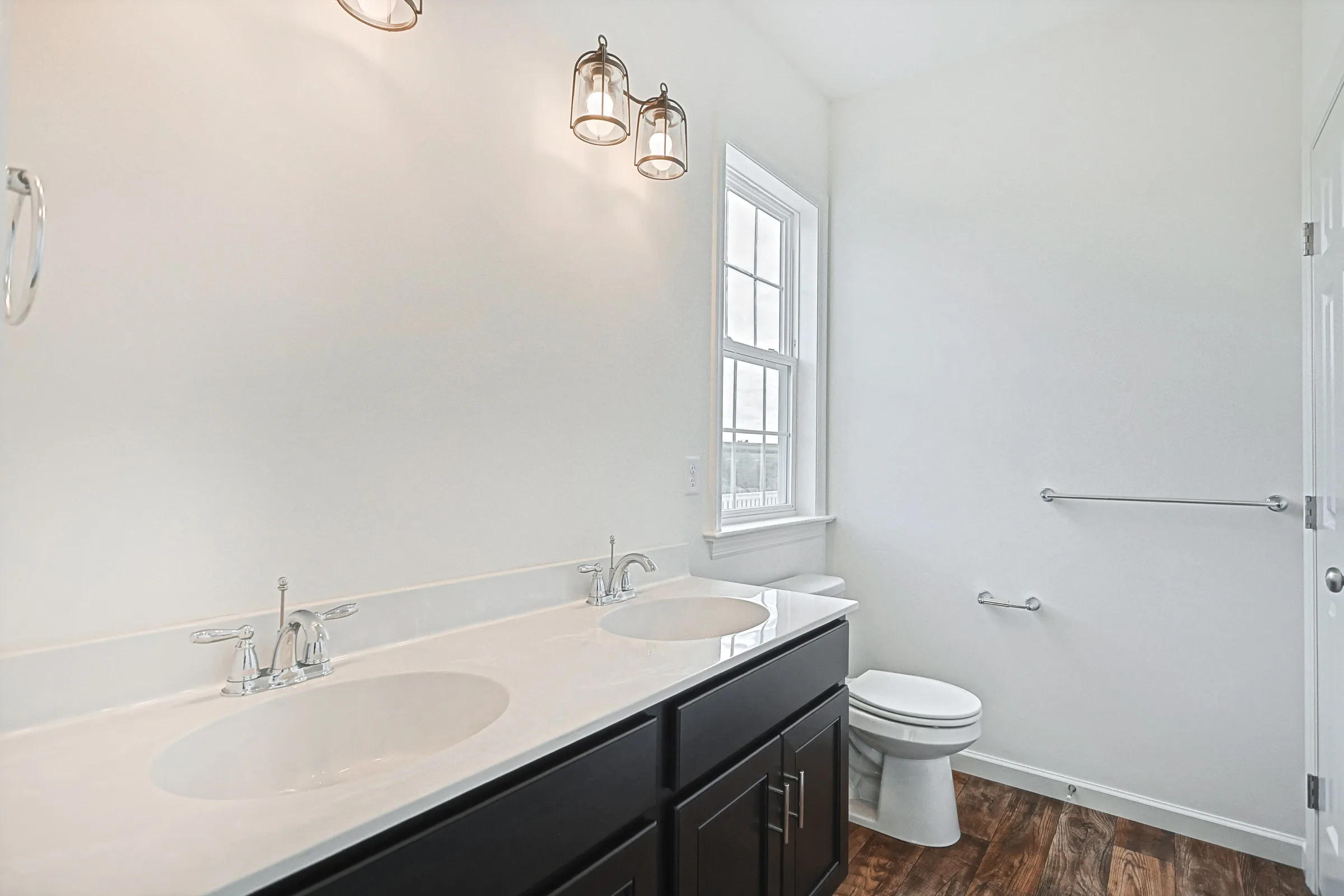 Modern bathroom with double sinks, dark cabinets, and a white countertop. There is a toilet near a window, wood-style flooring, and three glass pendant lights on the wall above the sinks.