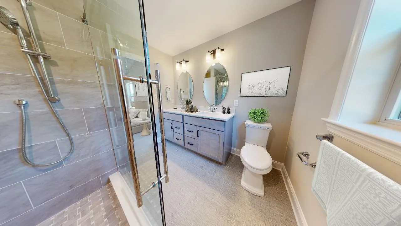 Modern bathroom with a glass-enclosed shower, double sink vanity with two mirrors, toilet, towel rack by a window, and neutral-colored walls and flooring. A potted plant and framed art add decorative touches.