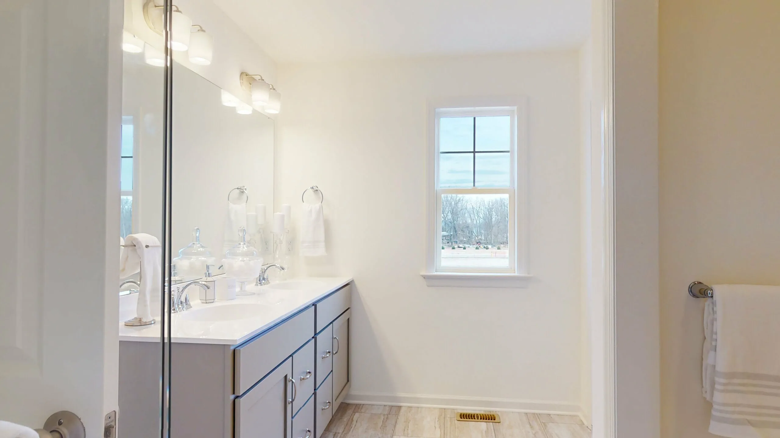 Bright, modern bathroom with a double-sink vanity, large mirror, and two wall lights. A window lets in natural light. Towels and decorative items are neatly arranged on the vanity and towel rack. Light wood floor.