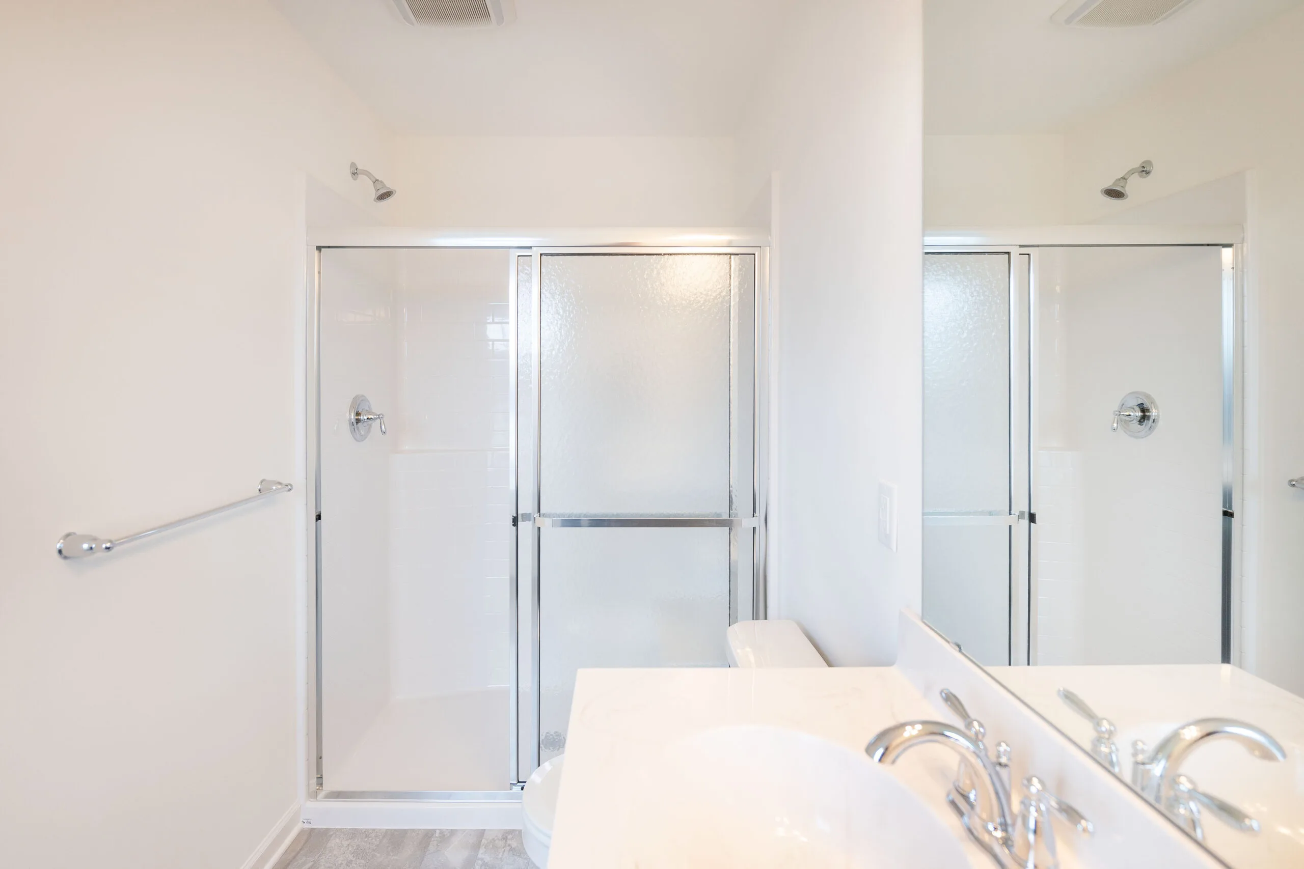 A bright, clean bathroom with a glass-enclosed shower, white walls, a chrome towel bar, and a white countertop with a silver faucet partially visible in the foreground.
