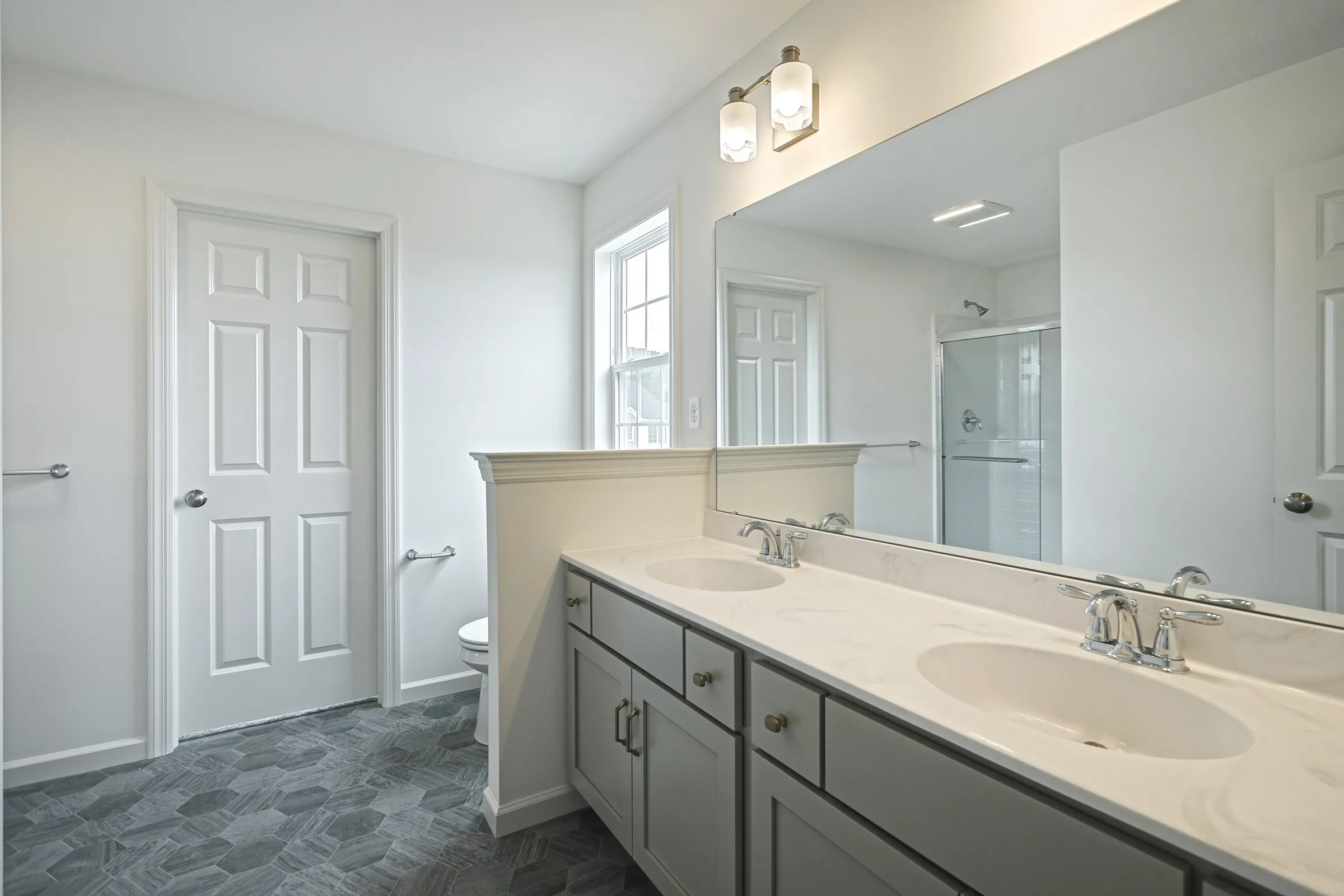 Modern bathroom with gray cabinets, double sinks, large mirror, and overhead lights. A toilet and shower with glass doors are visible in the background, along with two white doors and a window letting in natural light.