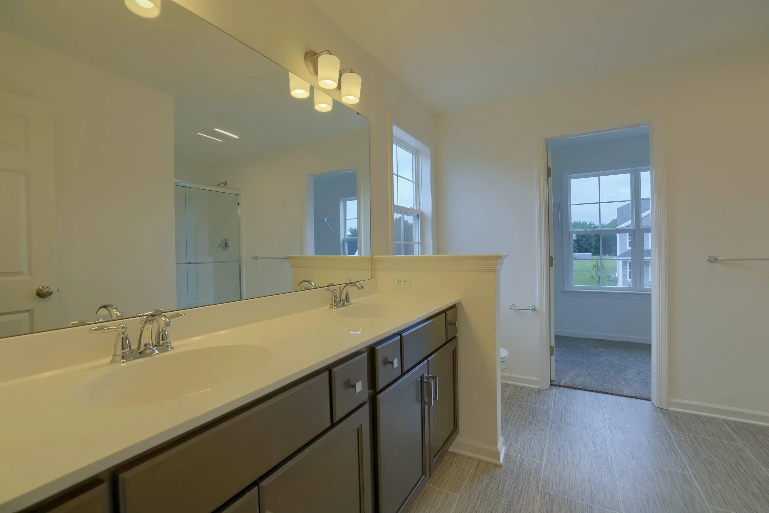 Modern bathroom with dual sinks and dark cabinets beneath a large mirror, soft lighting above, tiled floor, glass shower, and a window providing natural light. An open doorway leads to a carpeted room with windows.