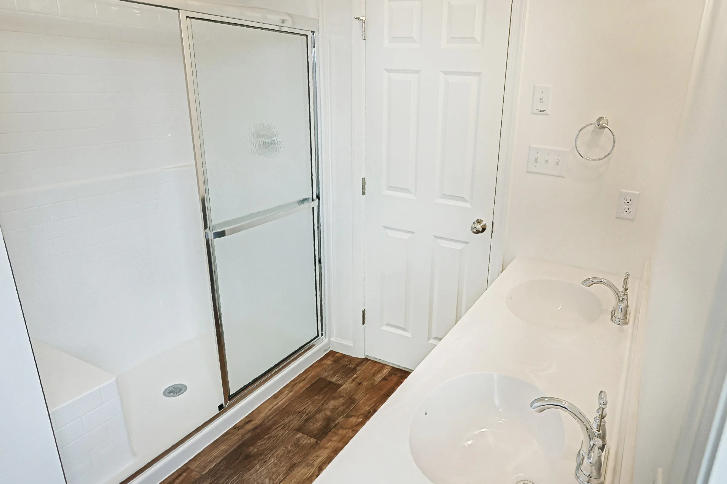 A bright bathroom with a glass shower enclosure, a white door, double sinks with chrome faucets, and a wood-style floor. The walls and counters are white, creating a clean, modern look.