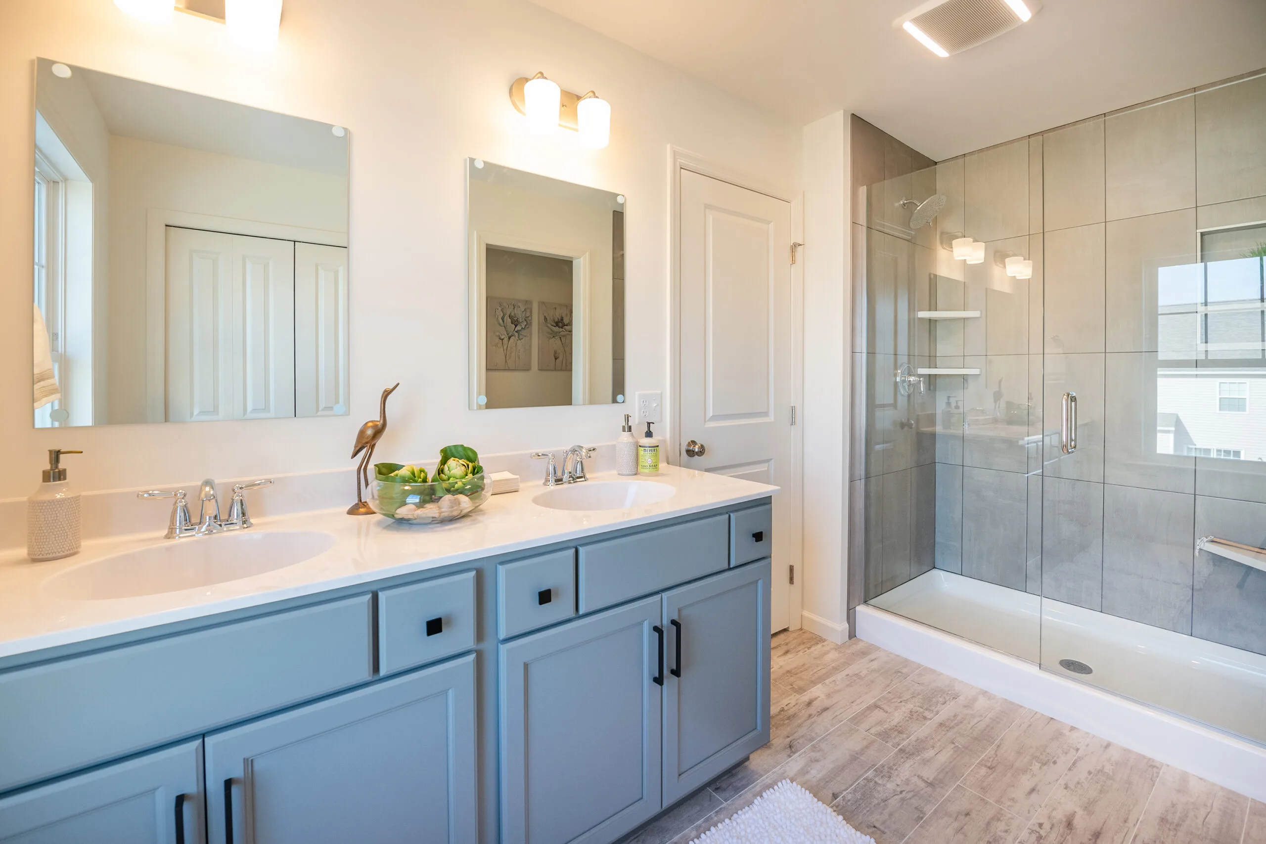 Modern bathroom with a double sink vanity, light blue cabinets, two mirrors, and a glass-enclosed shower. Bright lighting and light wood flooring create a clean, spacious look. Decorative items sit on the countertop.