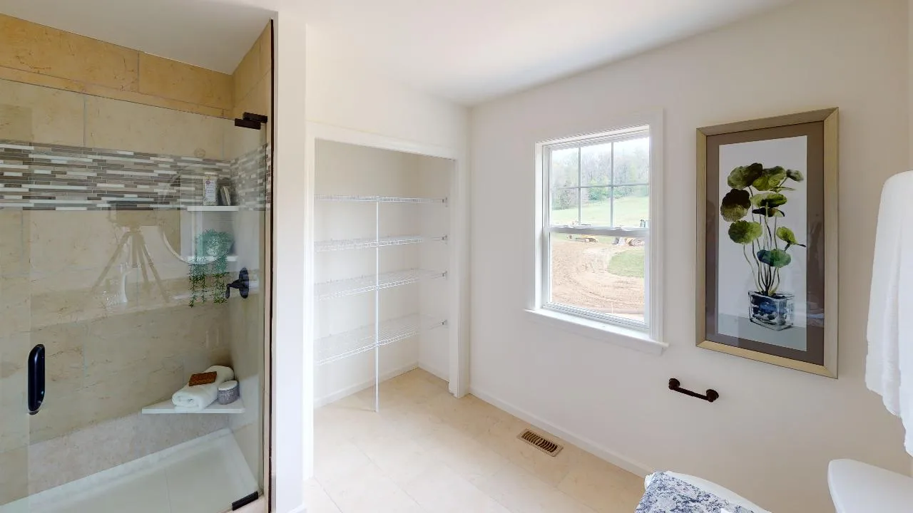 Bright bathroom with a glass shower, open shelving, a window with a scenic view, framed plant artwork on the wall, and beige tile flooring.