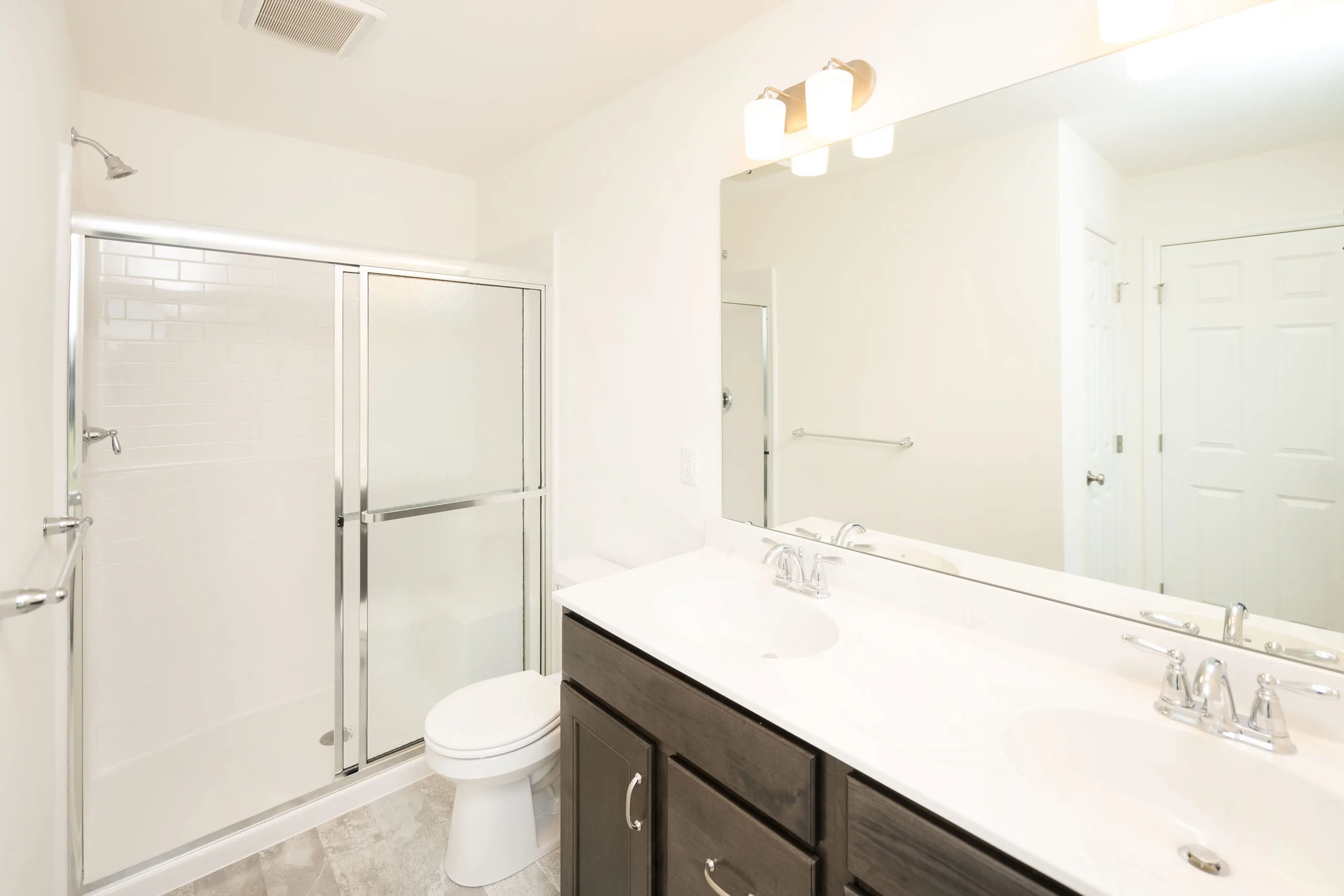 Bright, modern bathroom with a glass-enclosed shower, white toilet, dark wood vanity with dual sinks, large mirror, and overhead lighting. The room features light-colored walls and flooring.