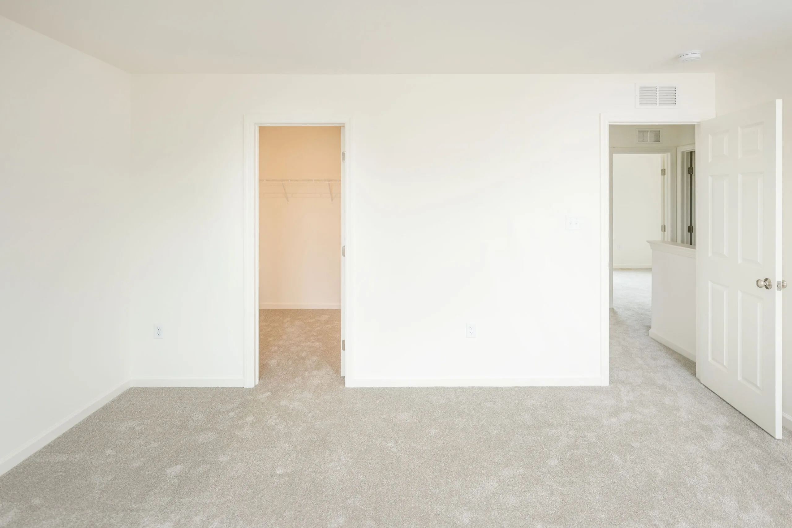 A bright, empty room with light-colored carpet, white walls, and two open doorways—one leading to a closet and the other to a hallway. The space has a clean, minimal, and freshly painted appearance.